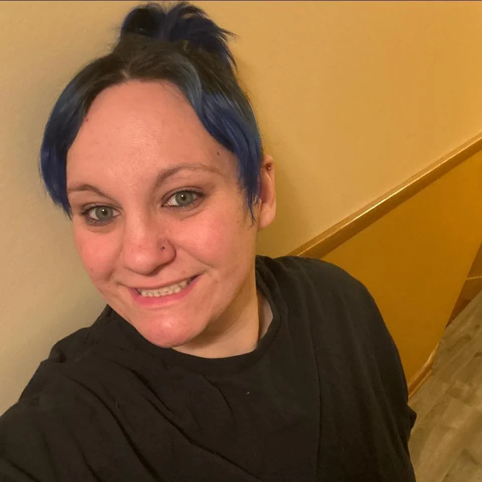 A woman with short blue and black hair, smiling, wearing a black shirt, standing in front of a beige wall with wooden staircase railing.
