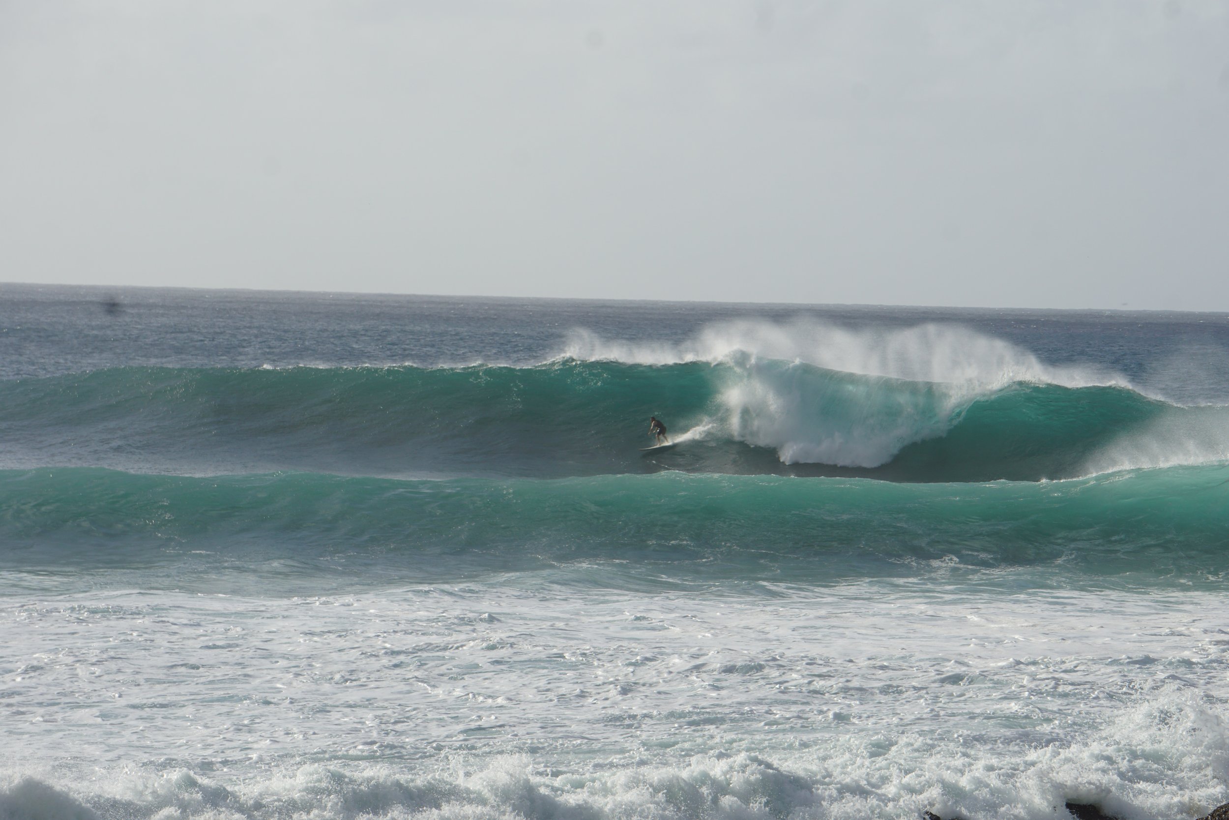 A surfer riding a large ocean wave near the shoreline with smaller waves in the foreground and another wave breaking in the background.