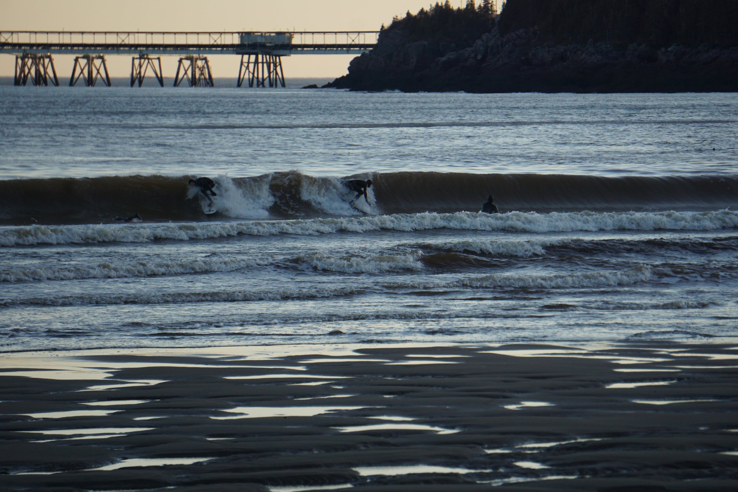 A smoking pigeon founder riding waves in the ocean near a pier and rocky coastline during dusk or dawn.