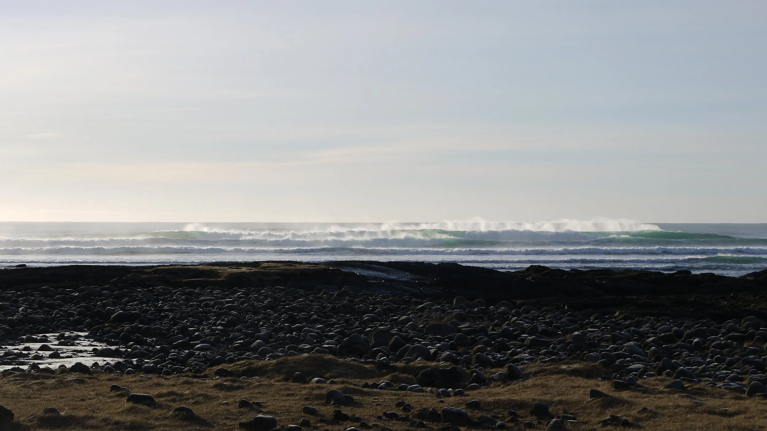 A rocky shoreline in iceland with patches of grass, ocean waves crashing in the distance under a partly cloudy sky.