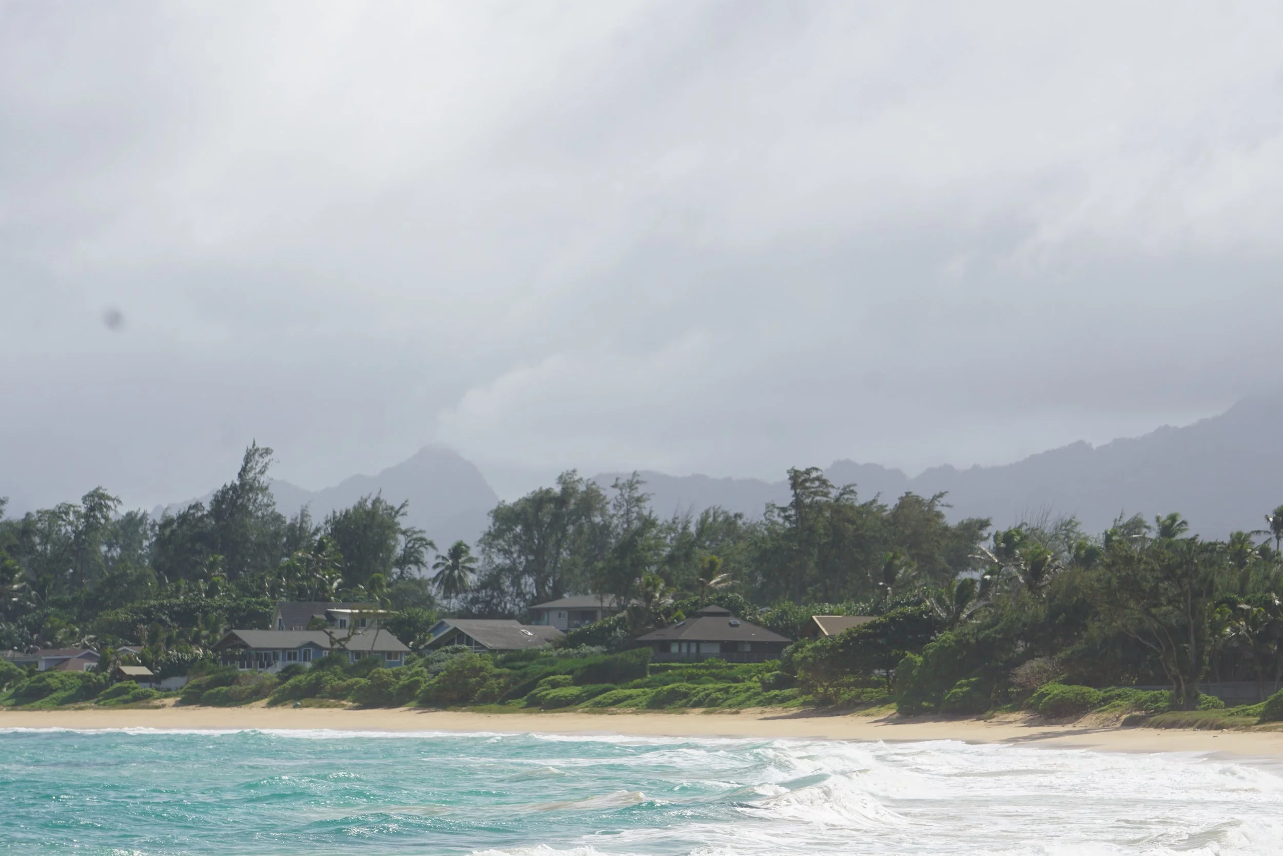 Beach with turquoise water, sandy shoreline, lush green trees, and houses, with mountains and cloudy sky in the background.