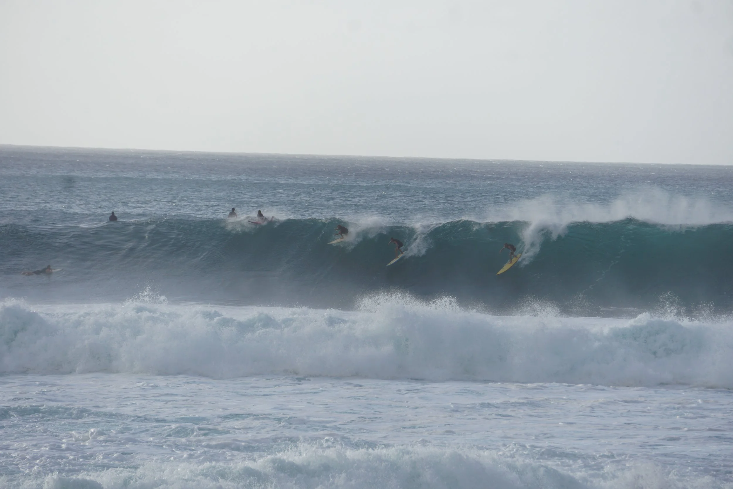 Multiple surfers riding large waves at the ocean, with some others waiting or paddling in the water.