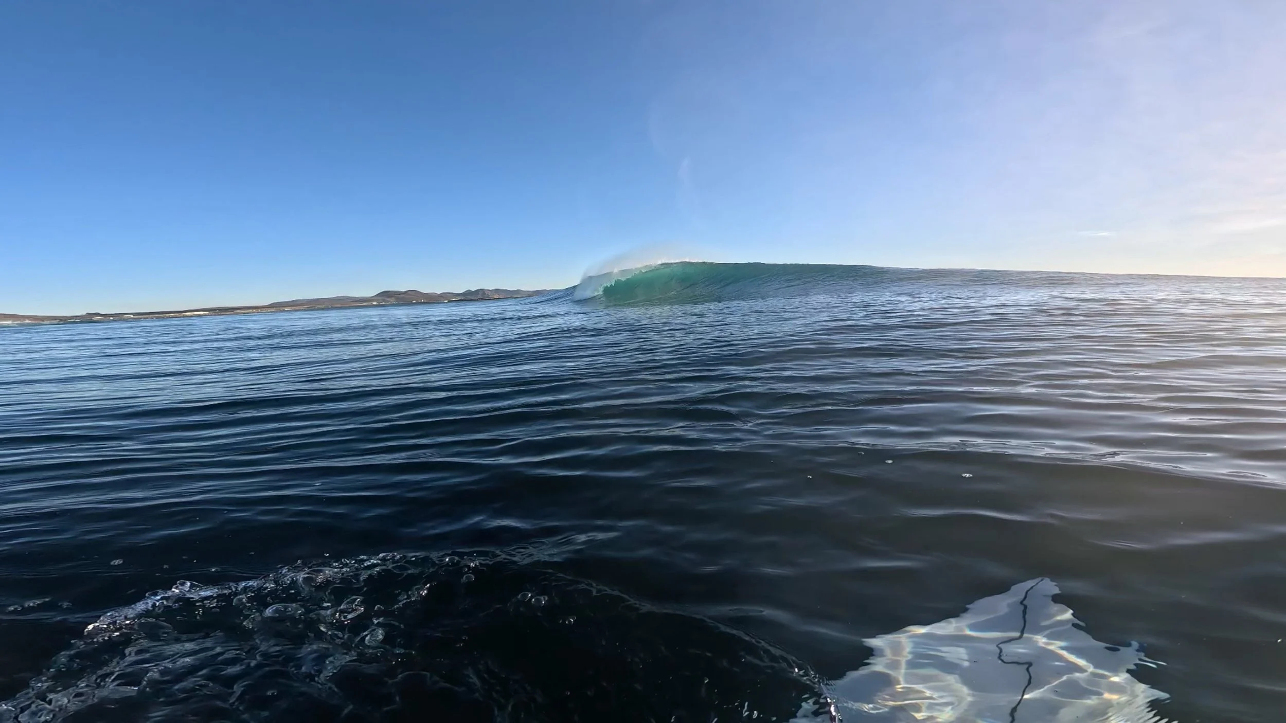 Ocean with waves, small landmass in the background, clear blue sky, and a large curling wave in the distance.