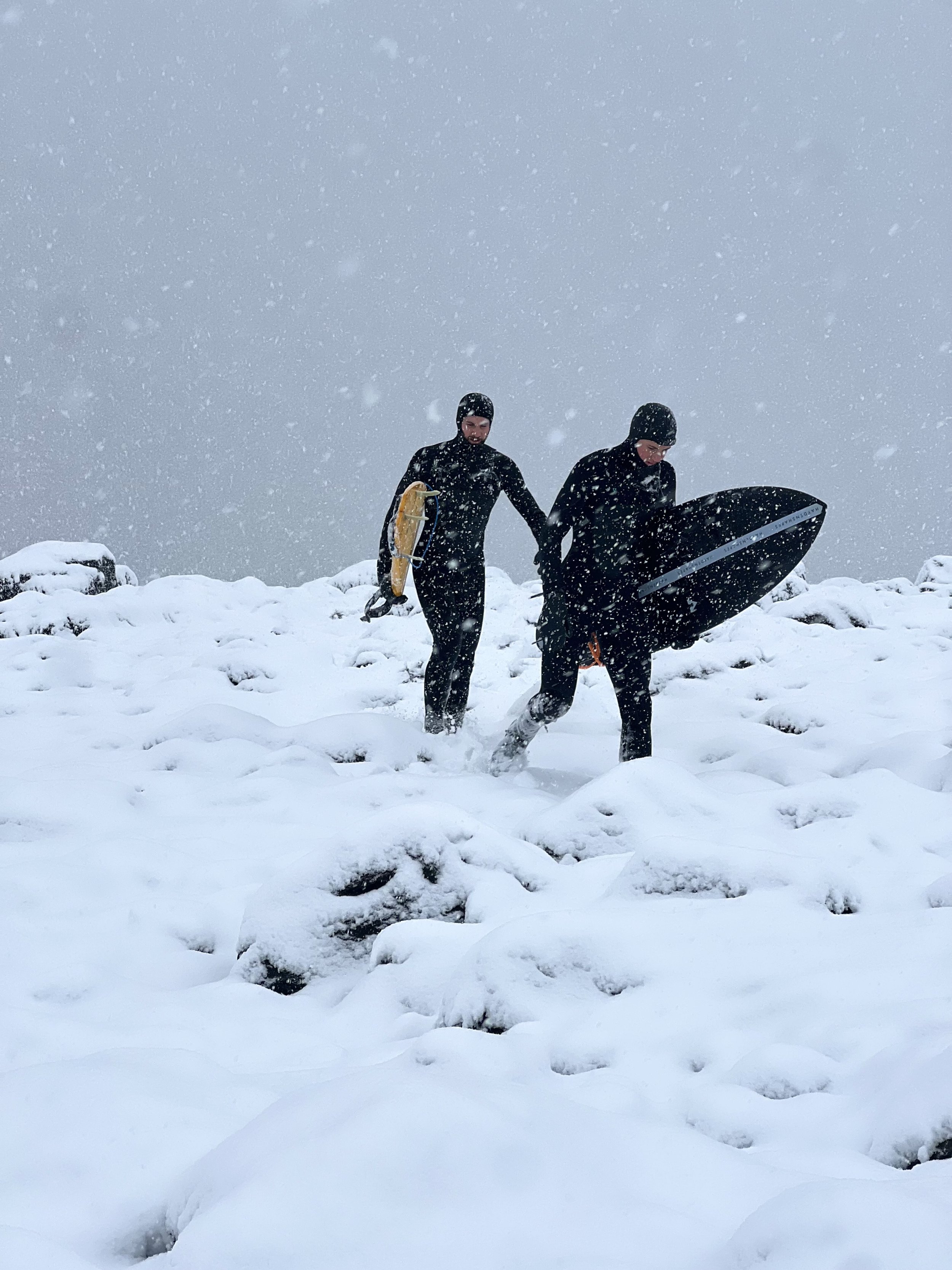 Two smokin pigeon founders  dressed in  wetsuits  carrying a surfboard and a yellow flotation device through a snowy, rocky landscape in a snowstorm in new brunswick canada