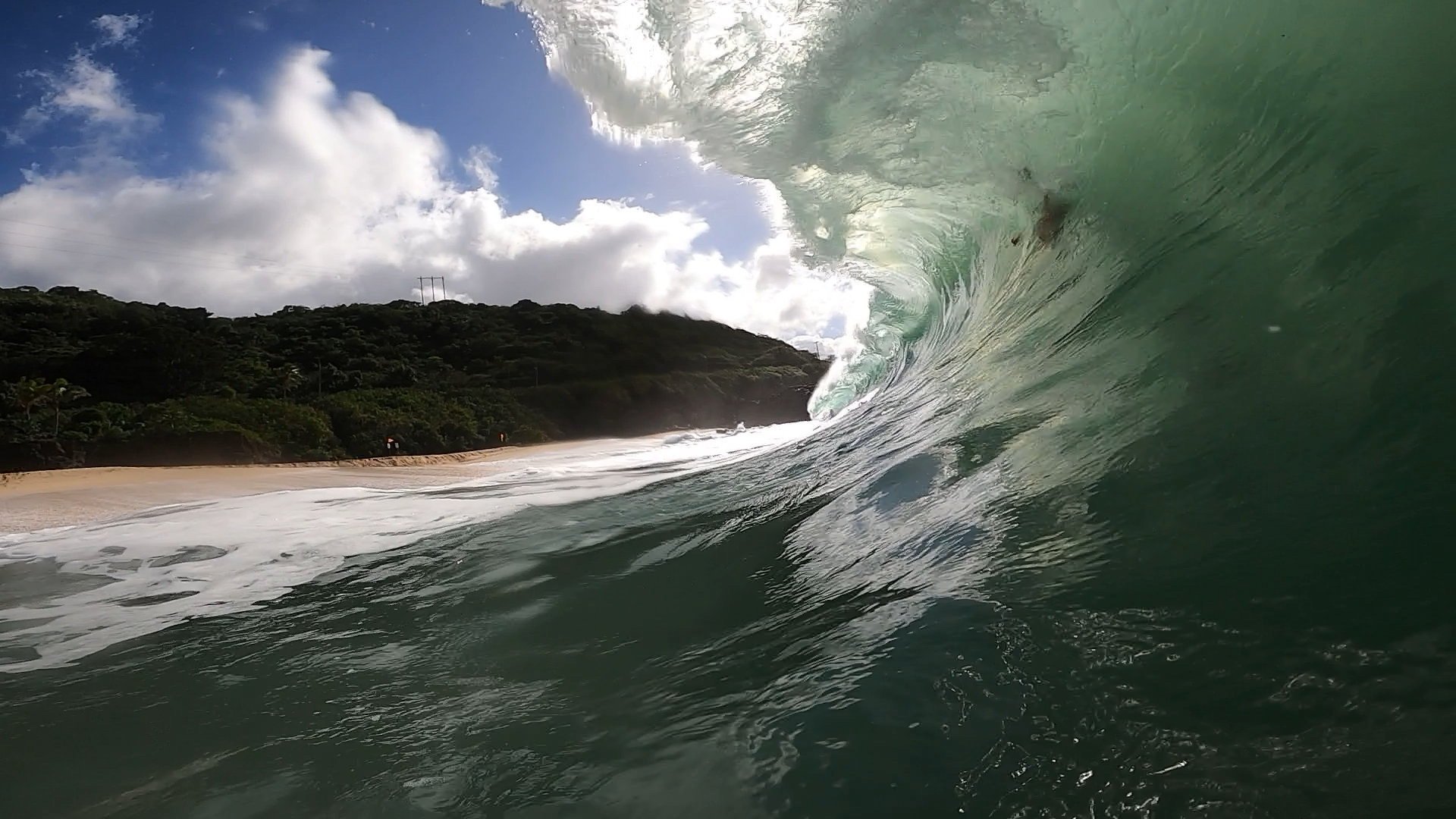 A large ocean wave crashing near a sandy beach with green hillside and cloudy sky in the background.