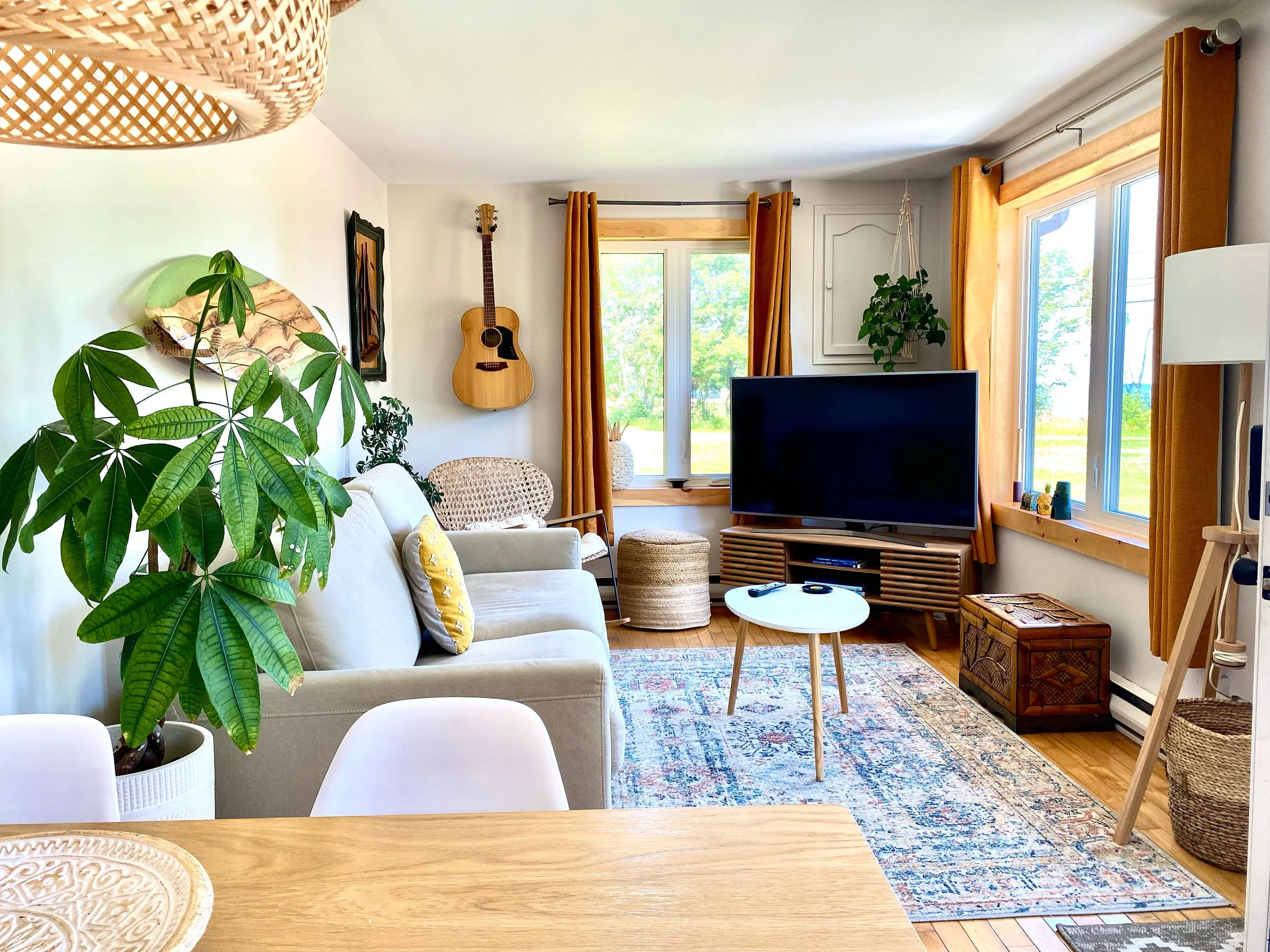 Living room with large window, cream sofa, potted plant, guitar on wall, flat-screen TV, patterned rug, and wooden furniture with mustard-colored curtains.