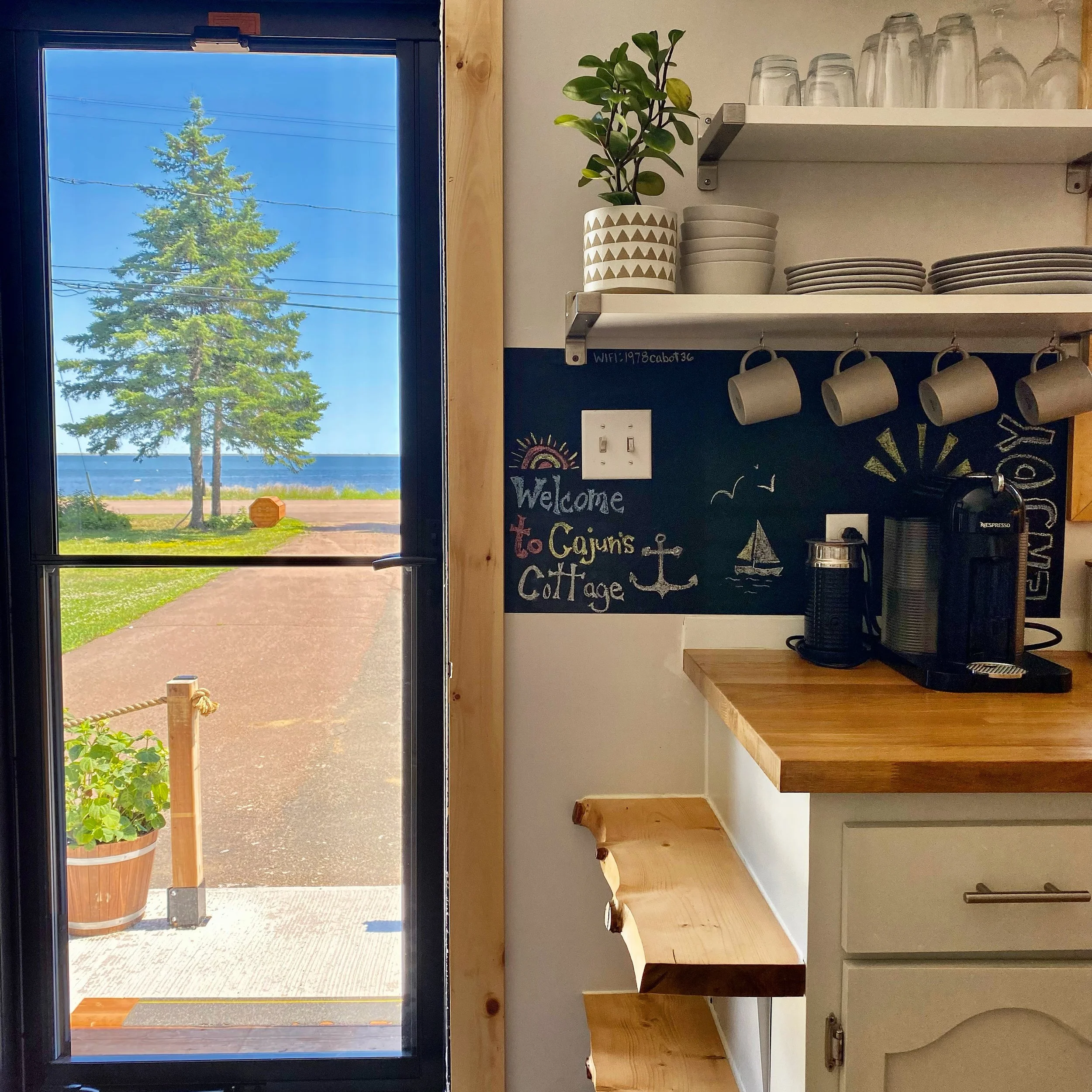 Inside a cottage kitchen with a view of a beach and ocean through a glass door. The kitchen has a wooden countertop, a coffee machine, and cups hanging from a rack. There are open shelves with plates, cups, and glasses, and a chalkboard with a welcoming message and seaside drawings.