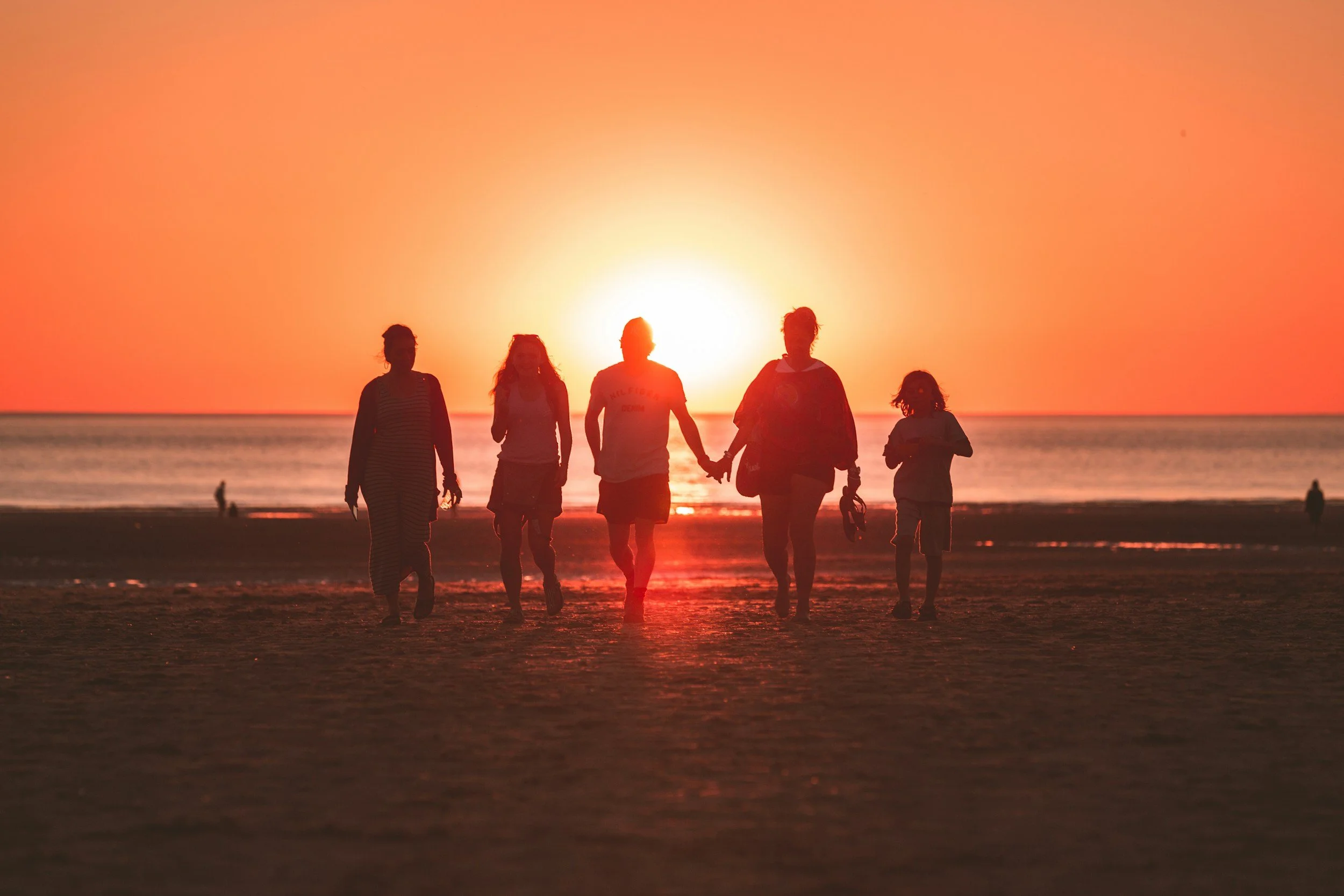 A group of five people walking on a beach at sunset, holding hands, with the sun low on the horizon and the ocean in the background.