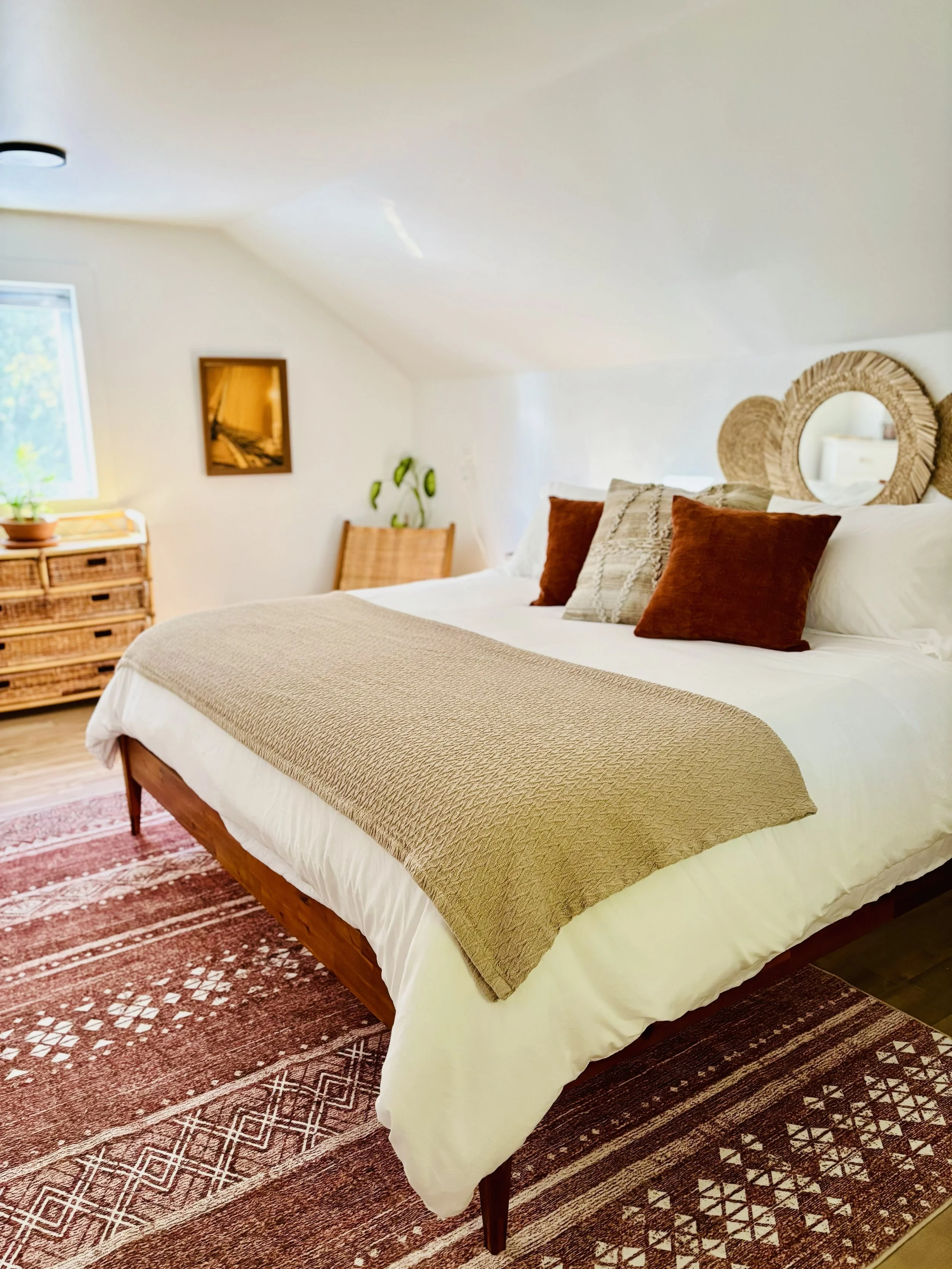 A cozy bedroom with a large bed covered in white bedding, a beige textured blanket, and decorative pillows in shades of brown and beige. There are two round mirrors with woven frames hanging above the bed. The room has a woven dresser near a window with a potted plant on it, and a framed picture on the wall. The floor features a patterned area rug with shades of red and cream.