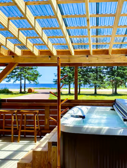 View from a covered porch overlooking a grassy area with trees and the ocean in the distance, featuring a hot tub and wooden chairs.