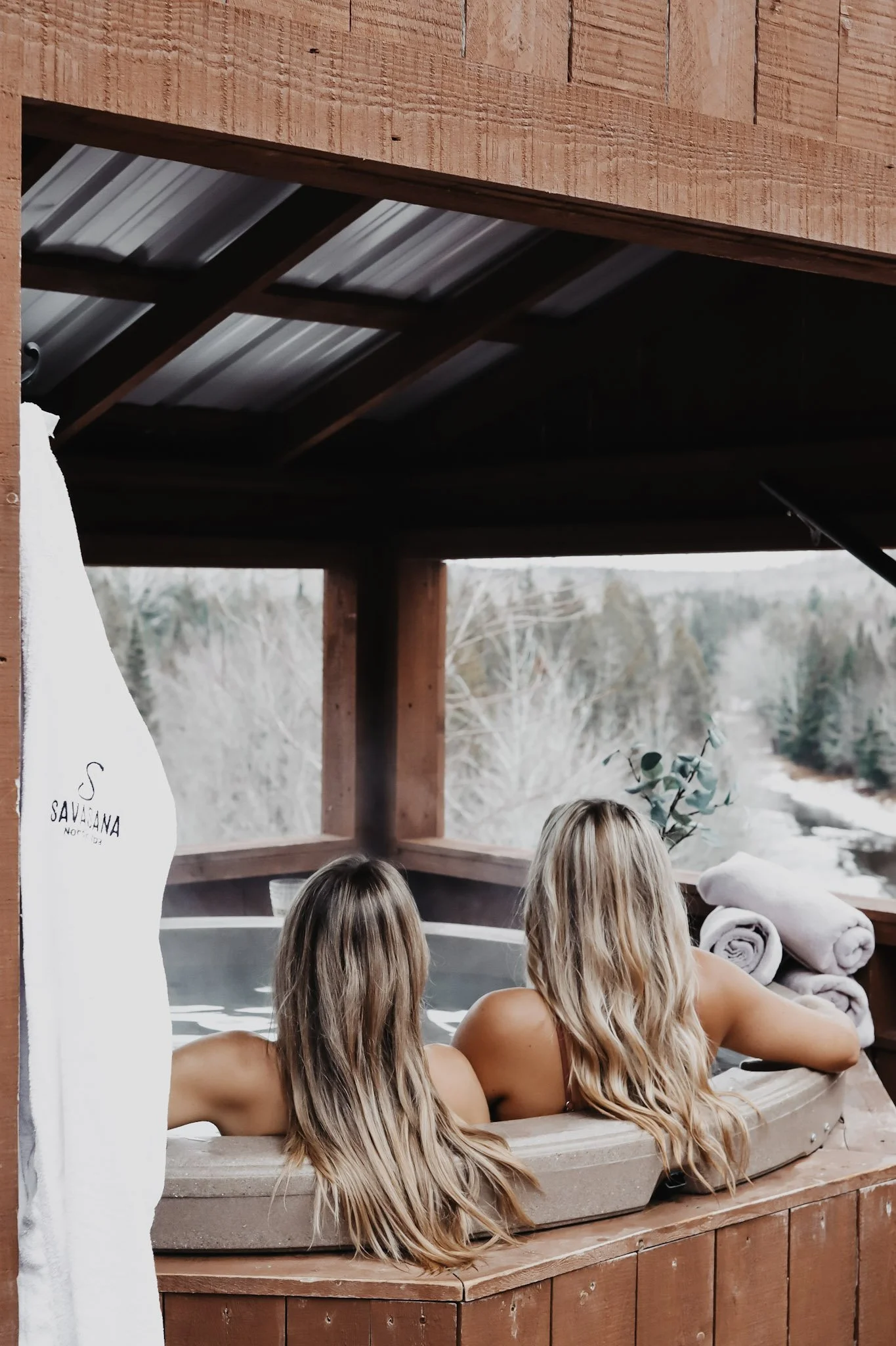 Two women sitting in an outdoor hot tub overlooking a wooded river landscape