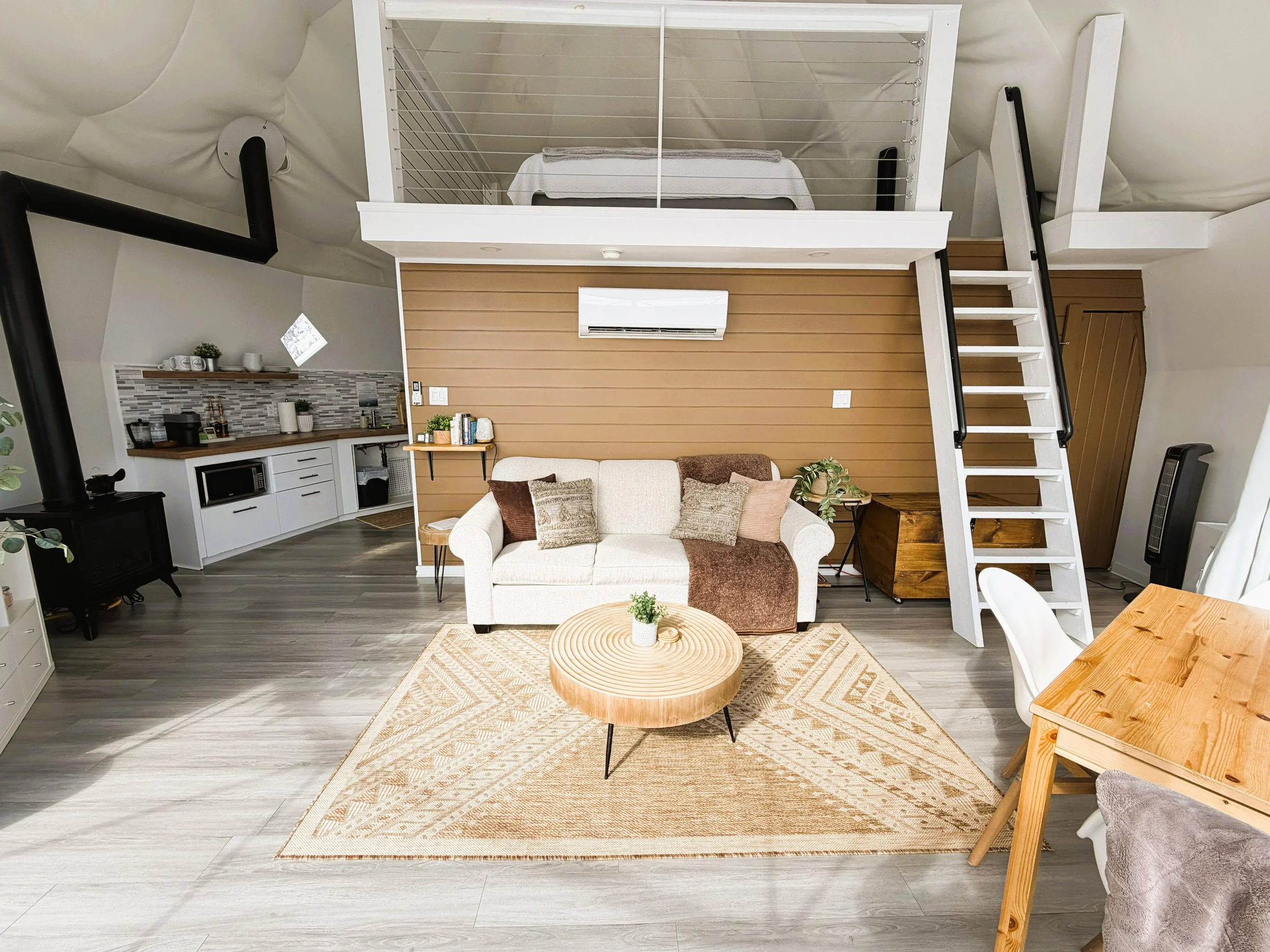 A cozy living room with a white sofa, beige cushions, a wooden coffee table, and a patterned rug. A lofted space above the living area features a bed and stairs with a black railing. The room has a wood-paneled accent wall, a small living space, and a visible kitchen area with white cabinets and a gray backsplash.