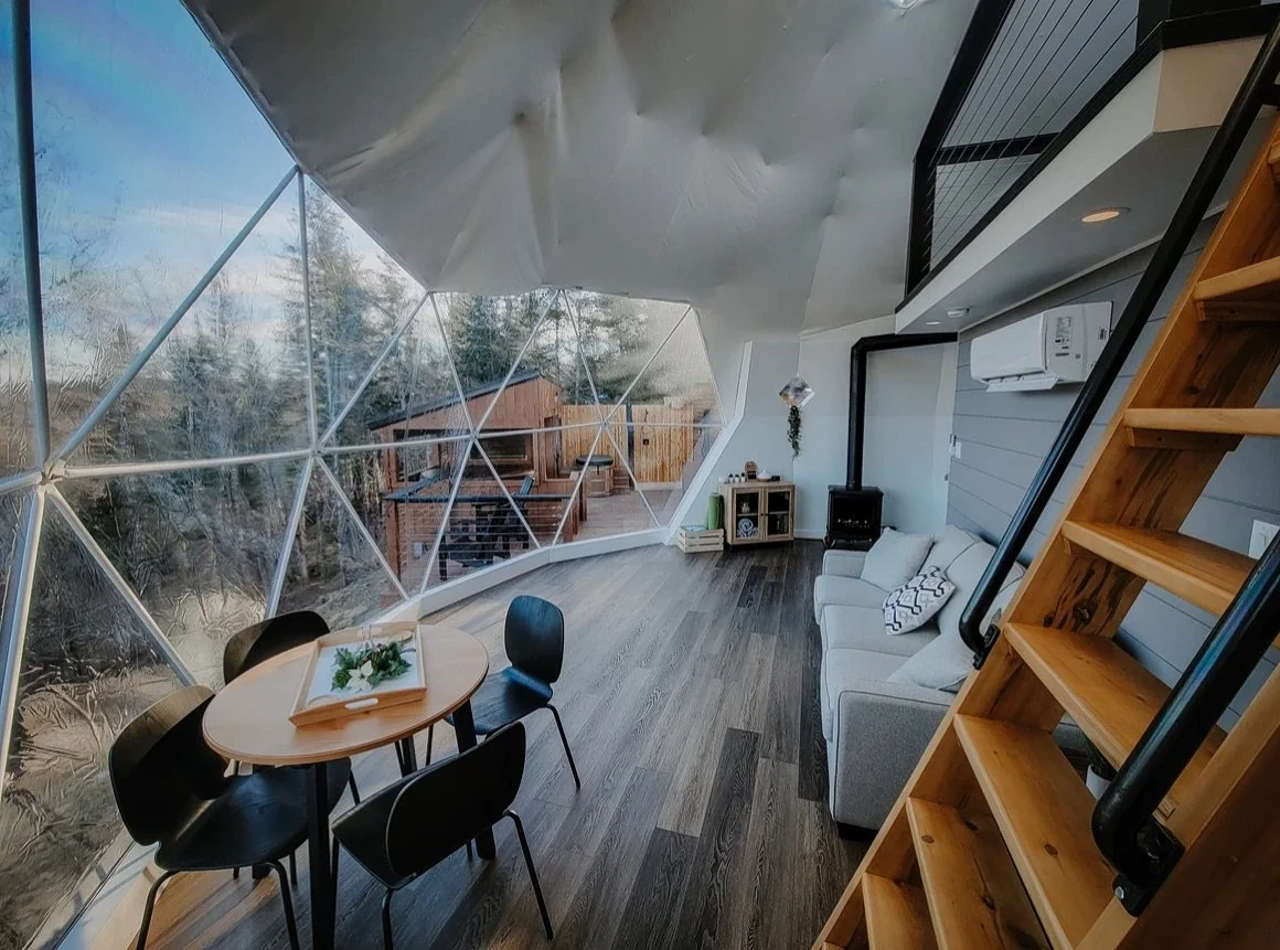 Interior of a modern geodesic dome with large glass windows, a small dining table with chairs, a beige sofa, and wooden stairs.