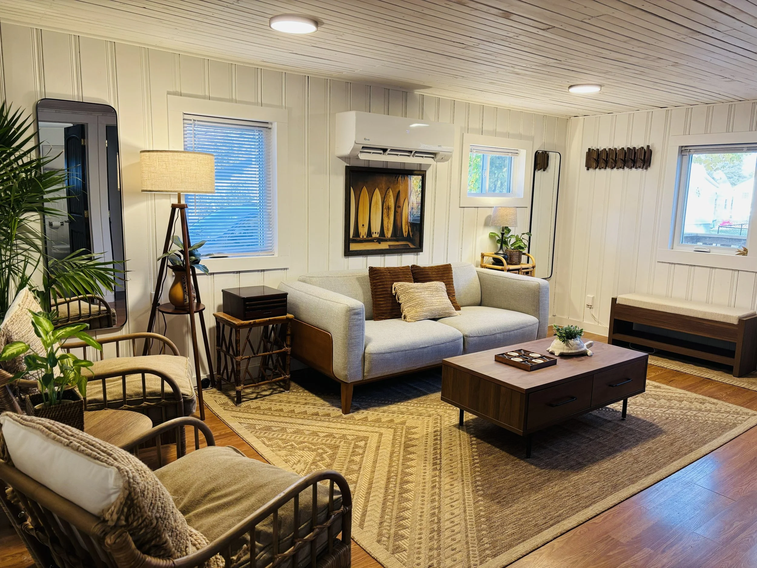 Living room with beige couch, wooden coffee table, wicker chairs, indoor plants, wall art, multiple windows, and hardwood floor.
