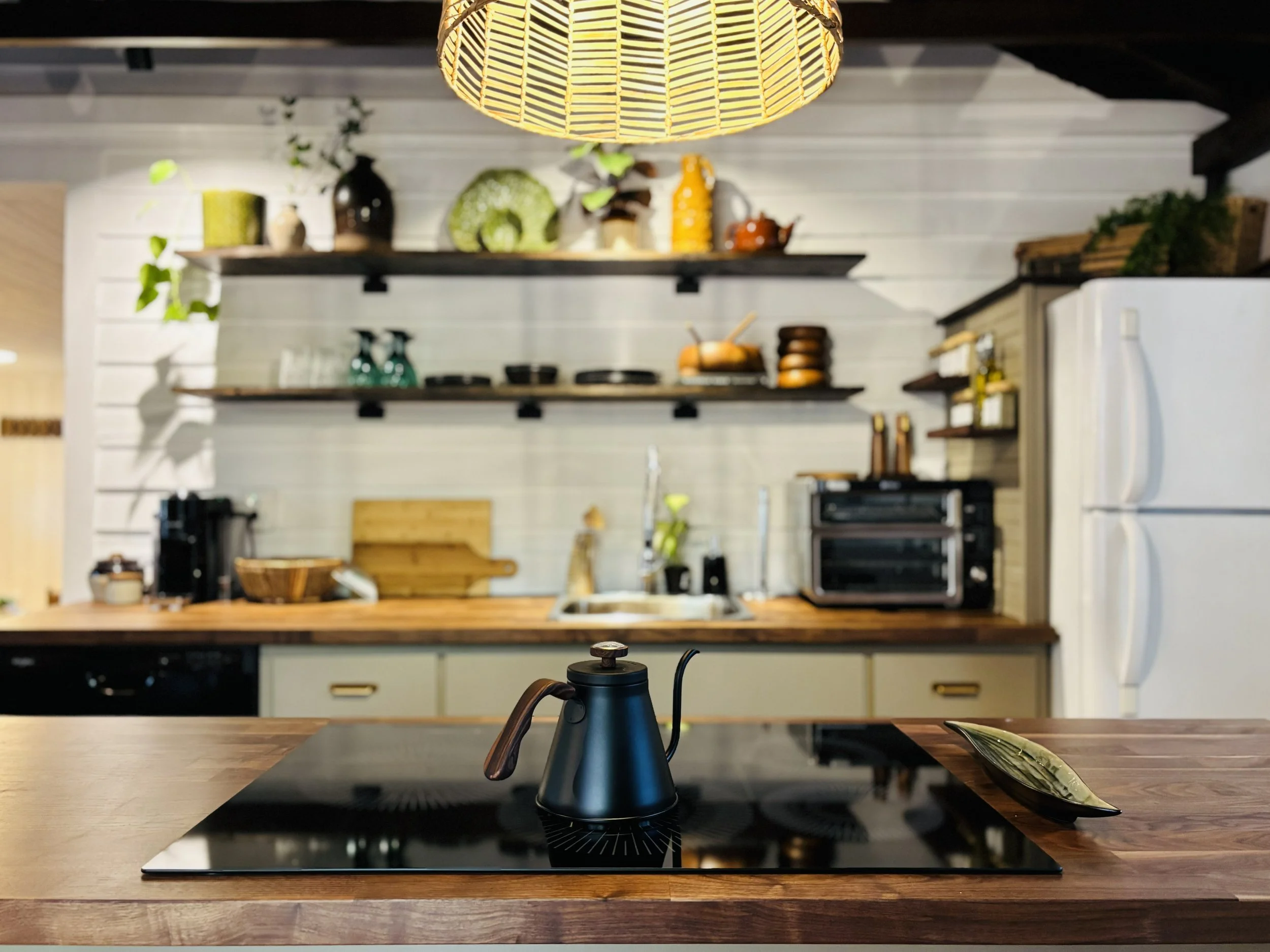 A modern kitchen with a wooden countertop, black stovetop with a kettle on it, and shelves with various decorative items and kitchenware in the background, including a white refrigerator.