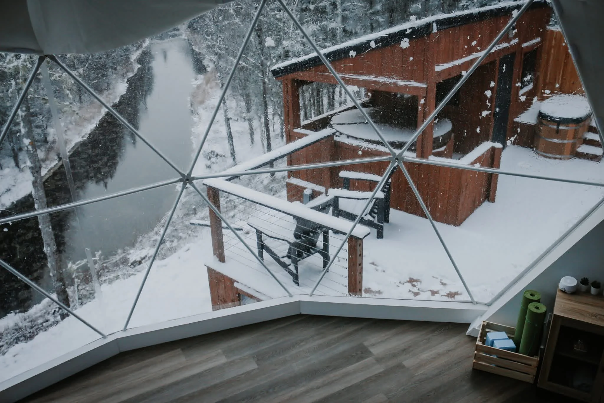 Snow-covered cabin balcony with a wooden hot tub and a small table, viewed through a large window with protective metal bars outside. Snow also covers the trees and ground in the surrounding landscape.