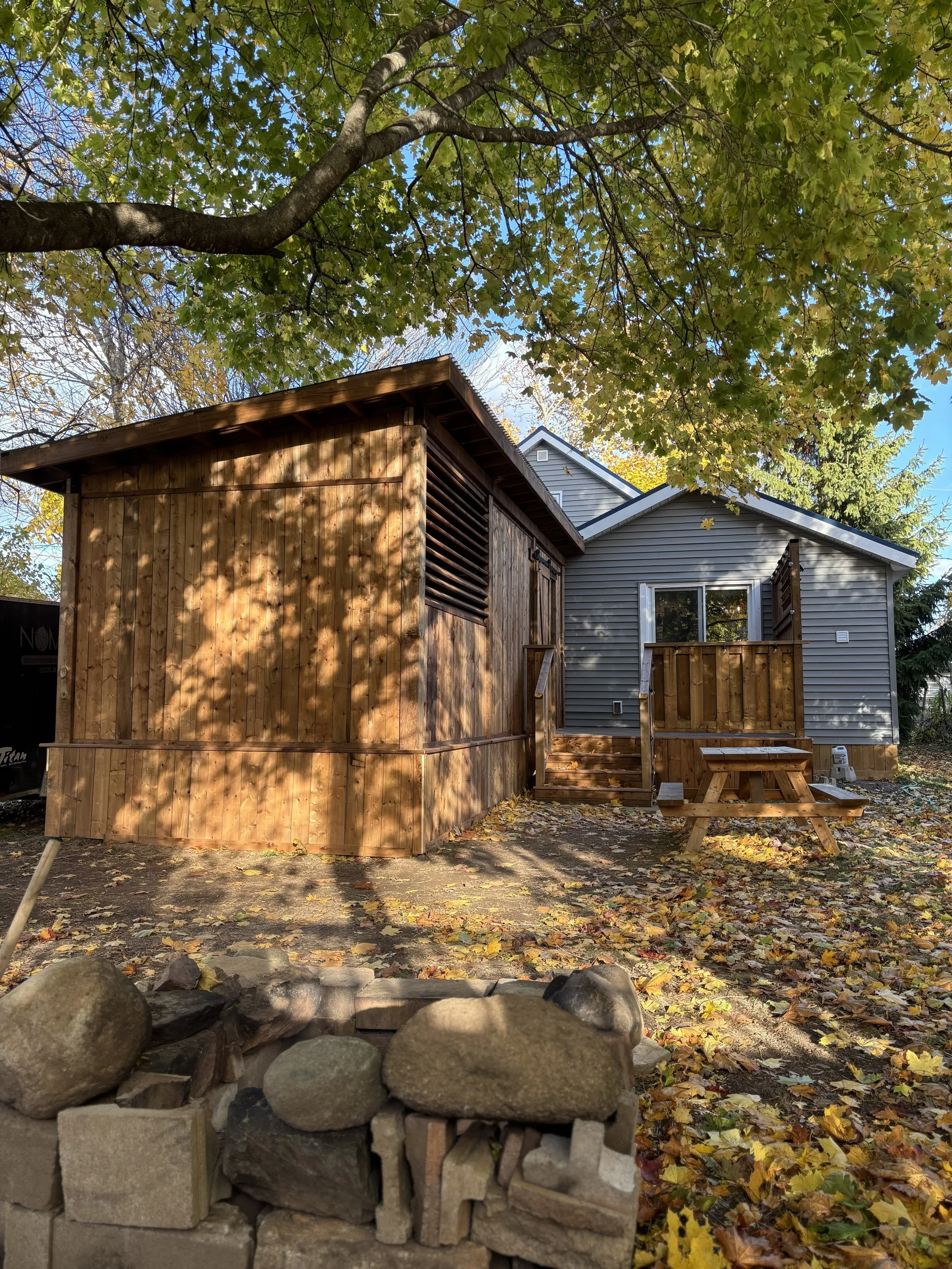 A house with gray siding and a deck with stairs, next to a small wooden structure, in a yard with fallen autumn leaves, large rocks, and a tree with green leaves overhead.