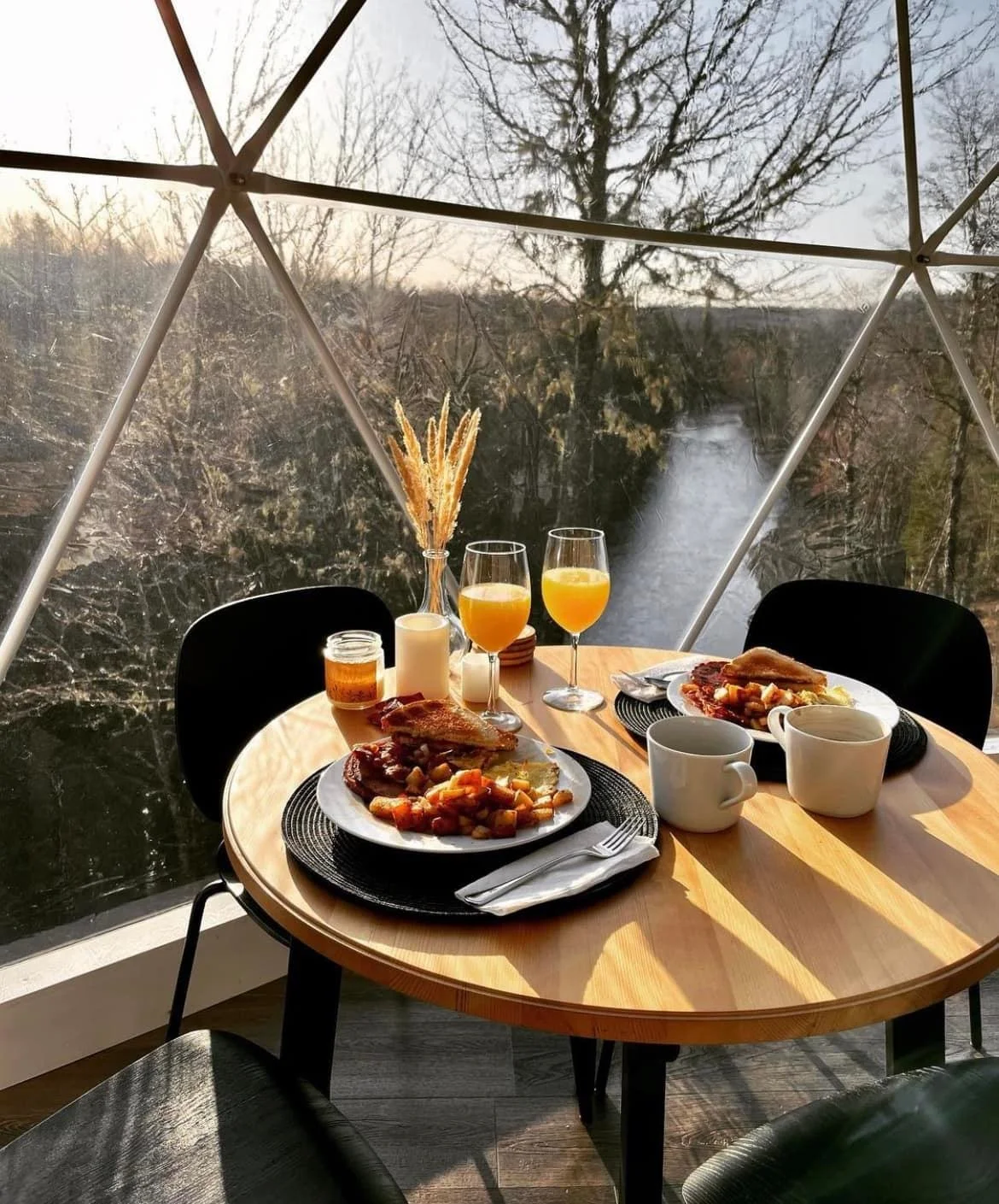 Breakfast table set with two plates of food, orange juice, coffee mugs, candles, and a vase with dried grass, inside a glass room overlooking a river and trees.
