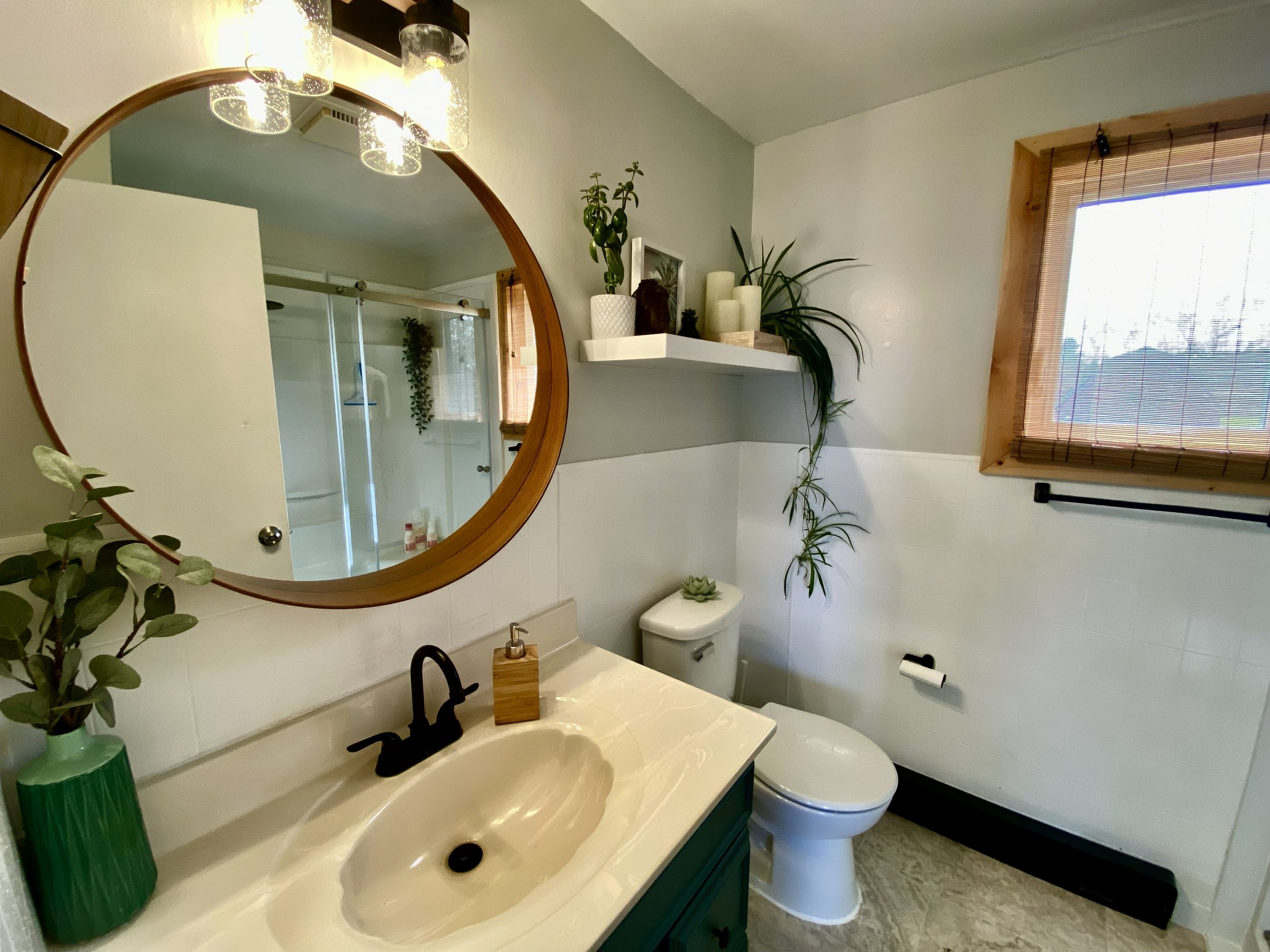 Bathroom with a large round mirror above a sink, black faucet, and green vase with leaves. Toilet with small plant on top, wall shelf with candles and plants, window with bamboo blinds, and a shower visible in the mirror reflection.