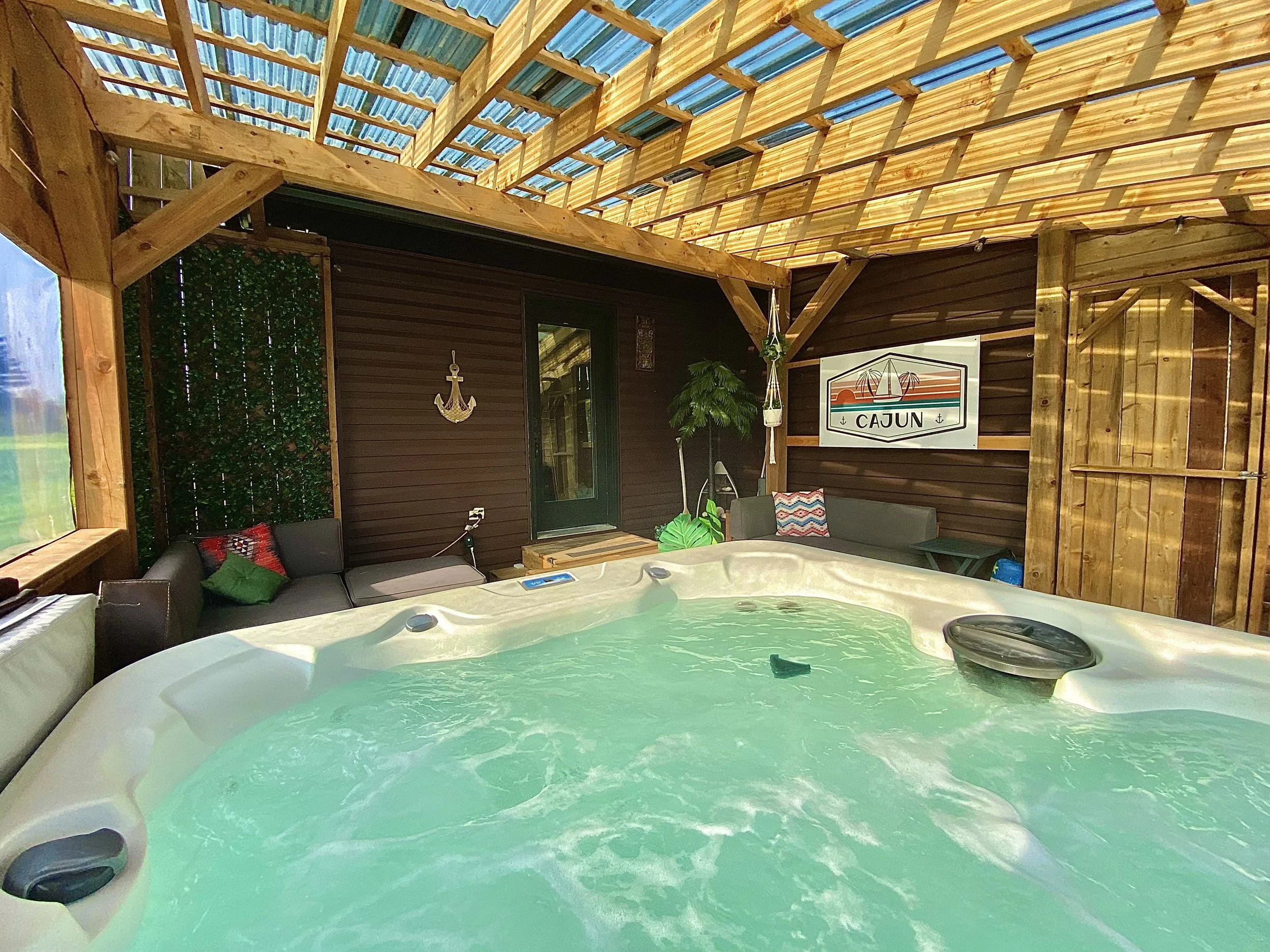 Indoor patio with a hot tub, shaded by a wooden roof, decorated with coastal-themed pillows, a large anchor wall hanging, a tropical plant, and a sign reading 'Cajun.'