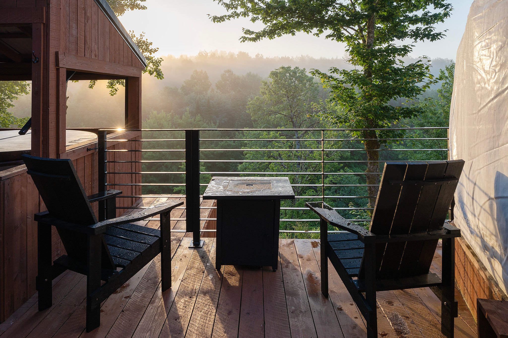 Sunrise over a forest view from a wooden balcony with two black chairs and a small table.