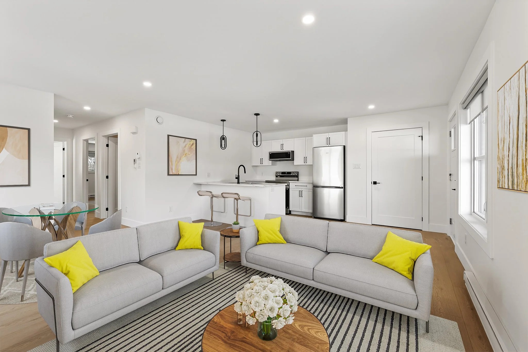 Open-plan living room with light gray sofas, yellow pillows, a round wooden coffee table with white flowers, a striped rug, a white kitchen with black fixtures, stainless steel appliances, pendant lights, and windows with white blinds.