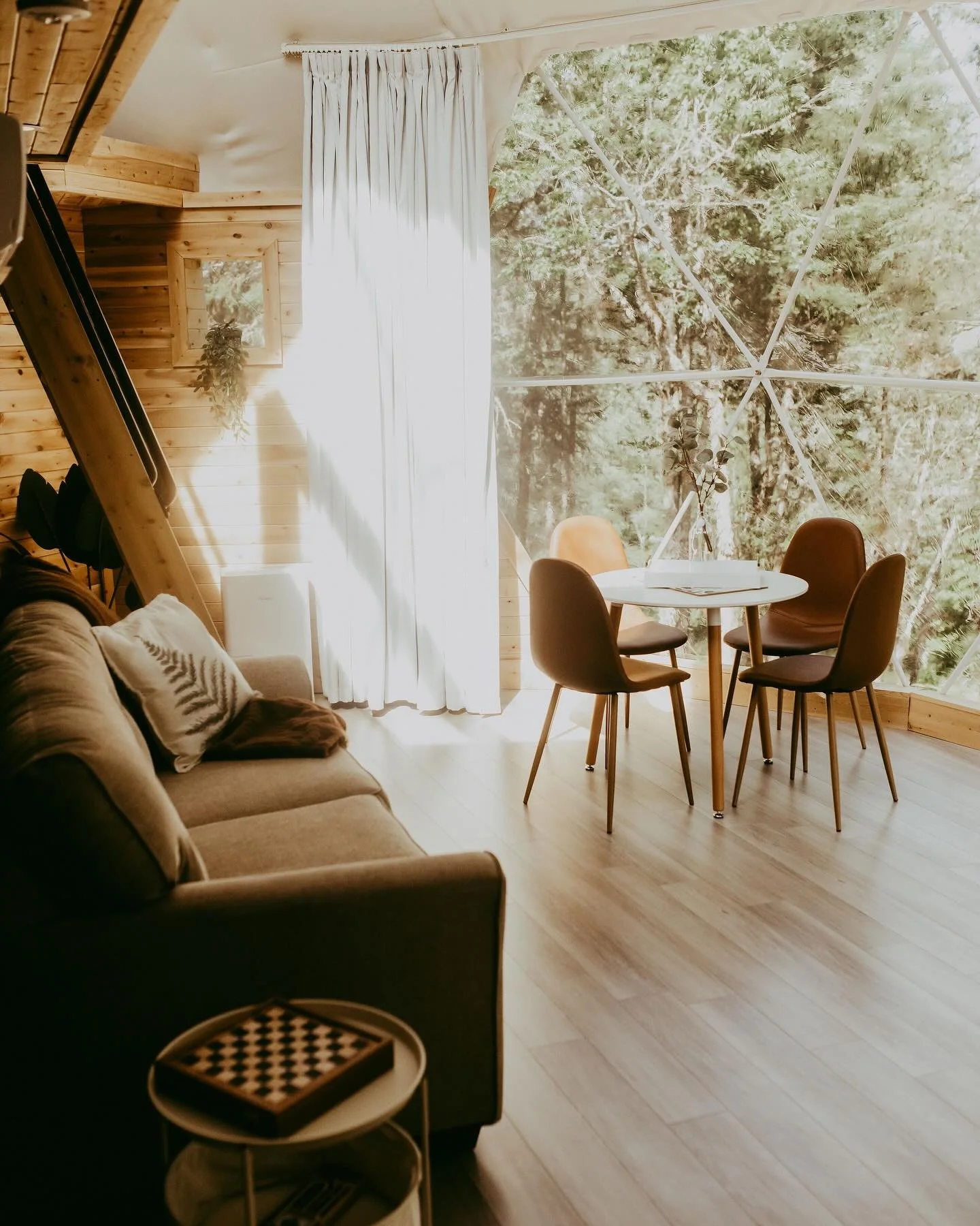 Sunlit cozy living space with a sofa, round dining table, four chairs, large window with a view of trees, and wooden interior elements.
