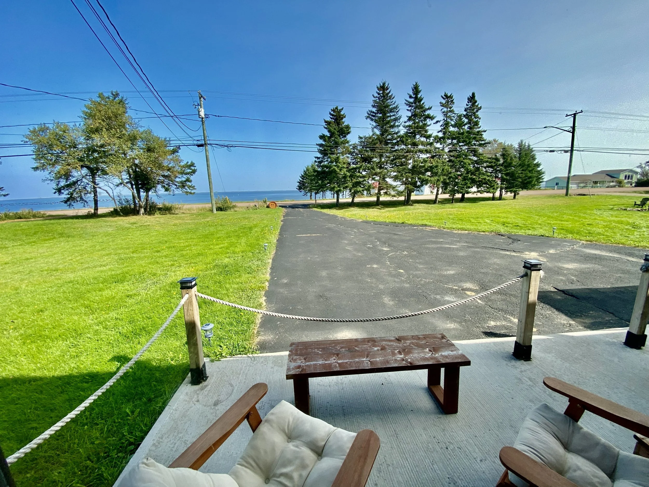 View from a porch overlooking a grassy area, trees, power lines, and a distant body of water on a sunny day.