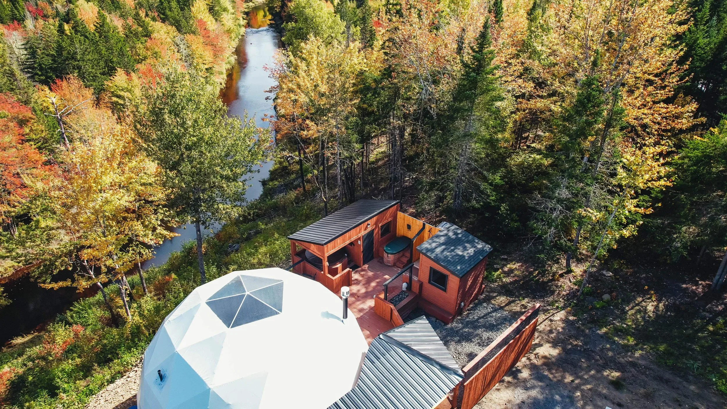 An aerial view of a small wooden cabin with a deck, hot tub, and outdoor seating area, surrounded by colorful autumn trees and next to a narrow river in a forest.