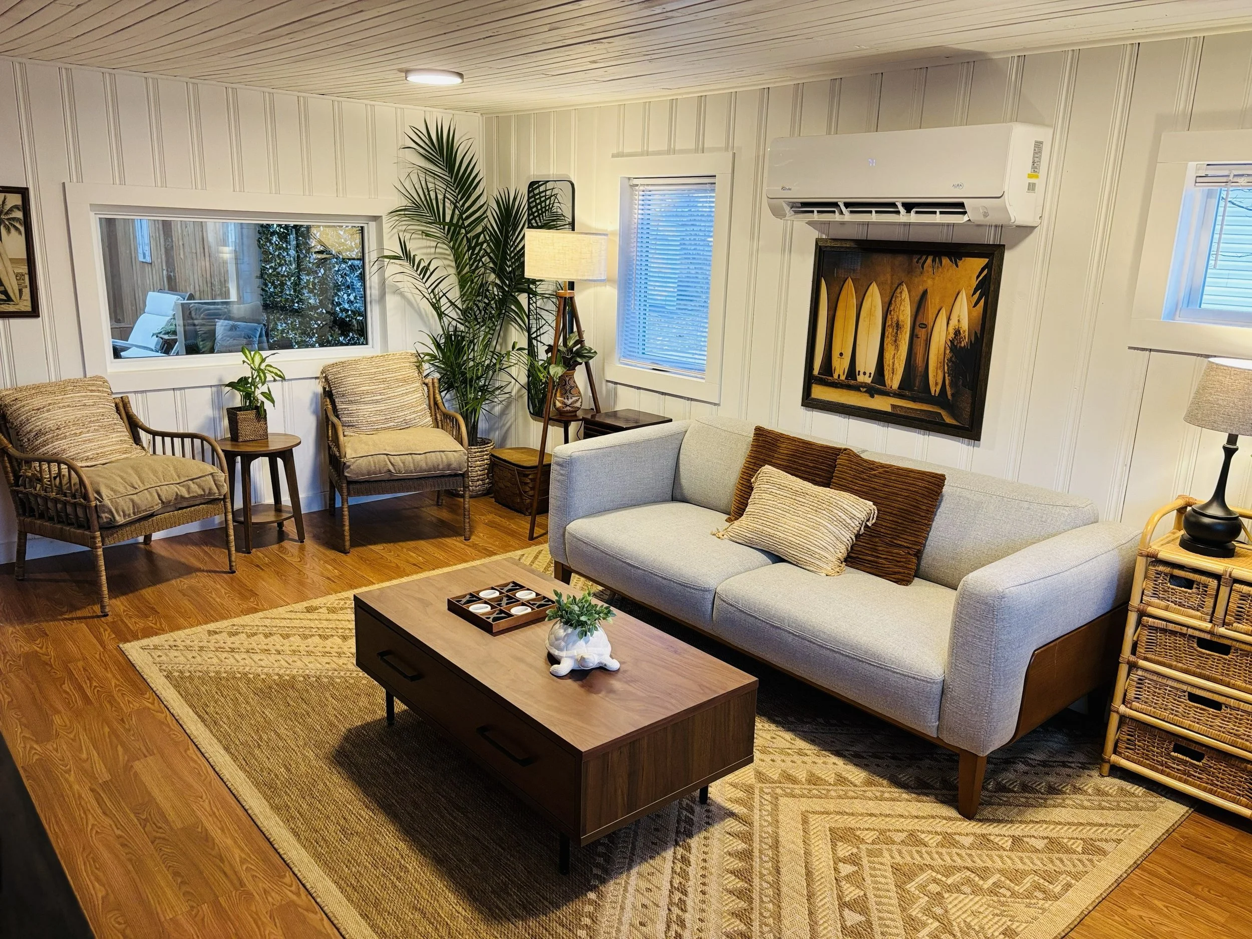 A cozy living room with white paneled walls, hardwood floors, and modern furnishings. There is a beige sofa with a throw pillow, a wooden coffee table with a decorative plant, and wicker chairs with cushions. Large windows and a wall-mounted air conditioner are visible, along with artwork and a floor lamp.