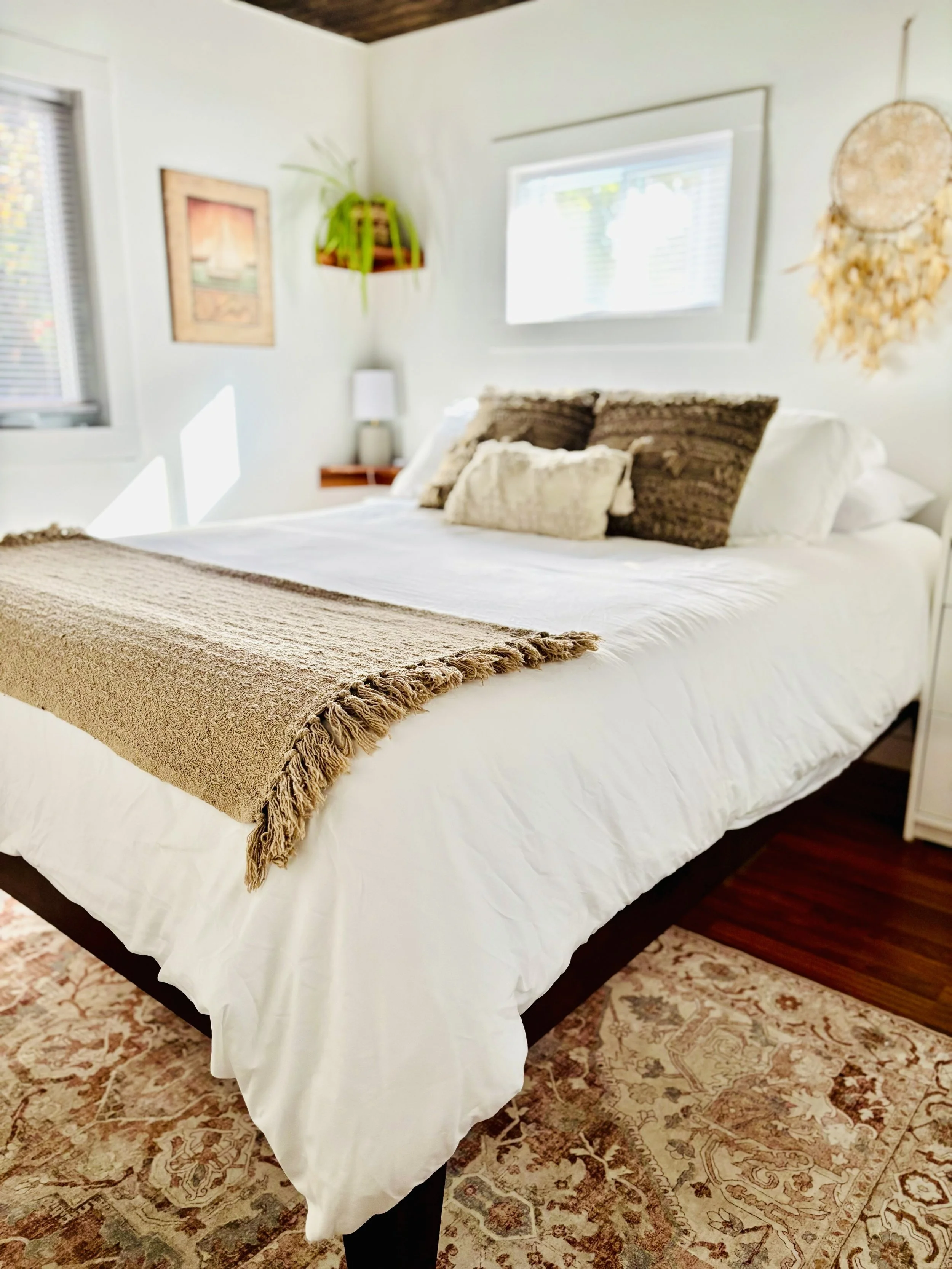 A neatly made bed with white linens, a tan throw blanket at the foot, and multiple decorative pillows. The room has white walls, a window with blinds, and decor including a wall hanging, a leafy plant on a shelf, a framed picture, and a dreamcatcher. There is a nightstand with a lamp next to the bed and a textured rug on the wooden floor.