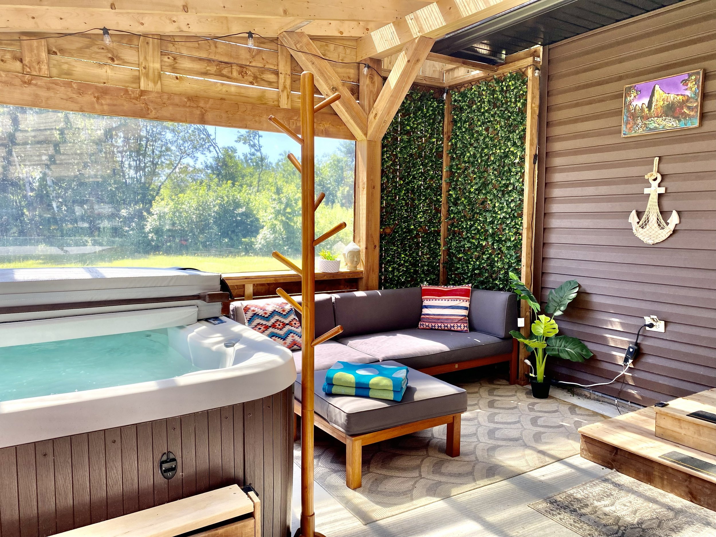 Cozy outdoor patio with hot tub, wooden furniture, potted plant, decorative pillows, wall art, and lush greenery.