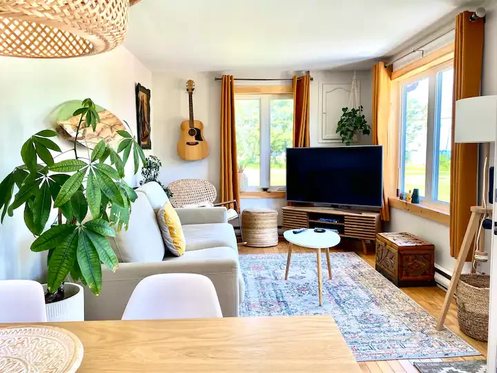 Bright living room with large windows, yellow curtains, white sofa with yellow and white pillows, potted plant, guitar on wall, flat-screen TV on wooden stand, woven ottomans, round coffee table, patterned rug, wooden cabinet, floor lamp, and a dining table in the foreground.