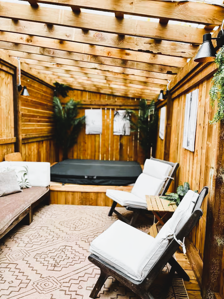 An outdoor covered patio with wooden walls and ceiling, featuring seating furniture, lush plants, and a hot tub at the back.