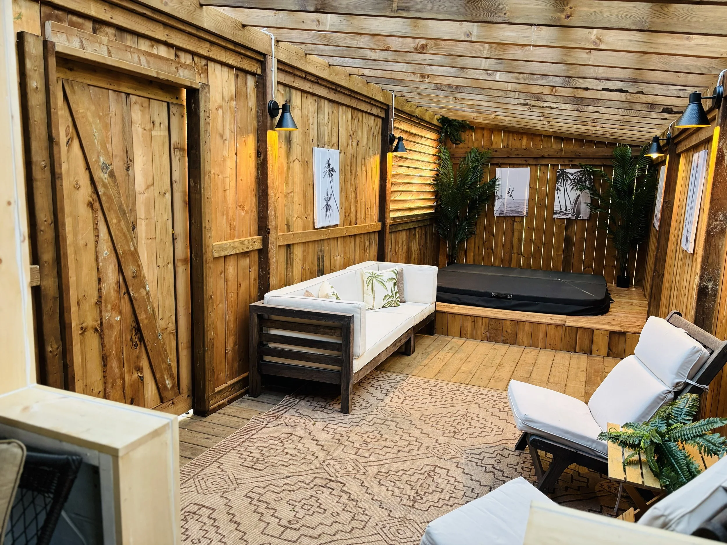 Interior of a wooden deck with tropical decor, including a white couch with pillows, a lounge chair, a hot tub, green plants, and framed coastal artwork.
