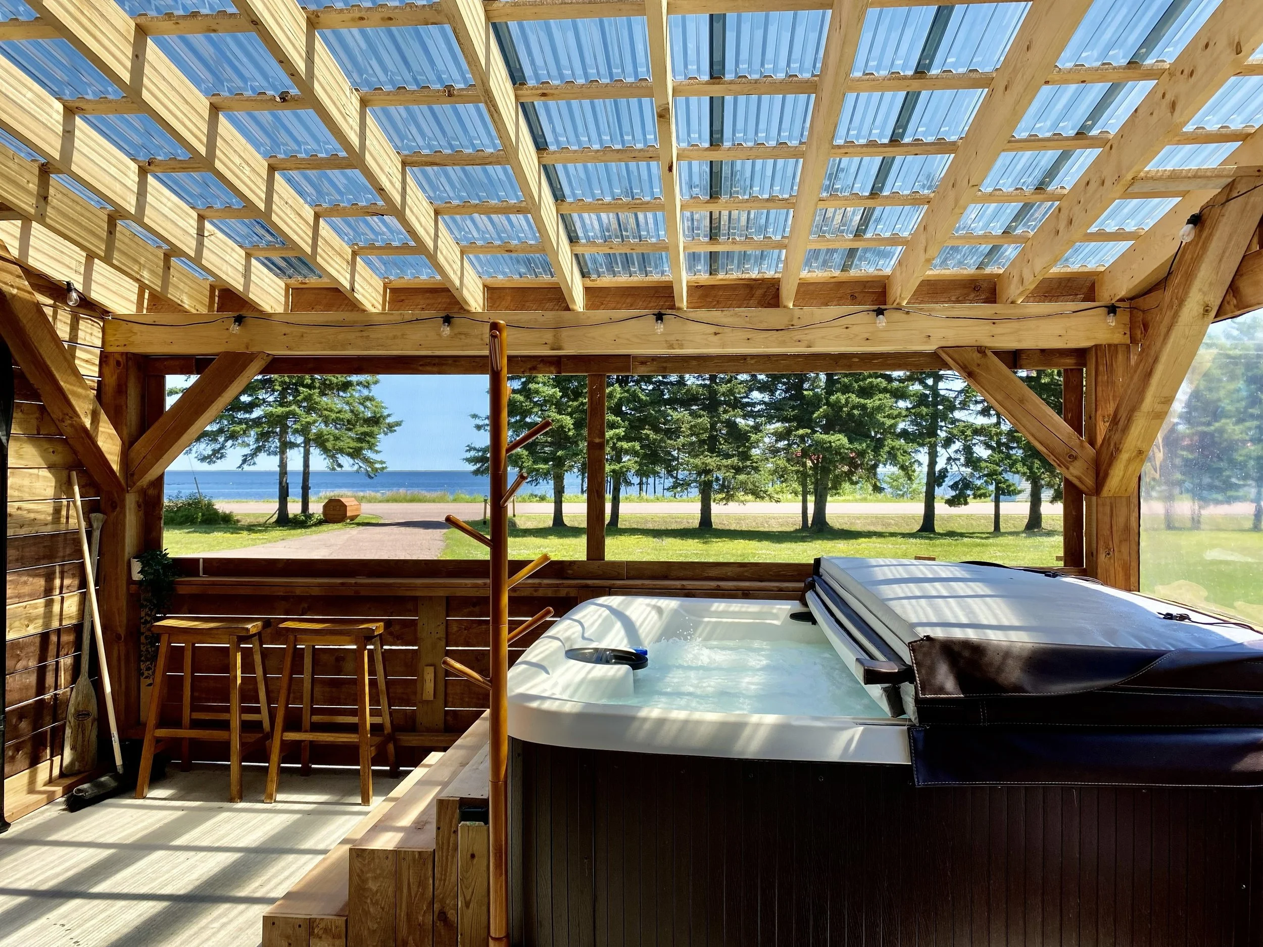 A covered outdoor space with a hot tub, overlooking a lake with trees in the background.