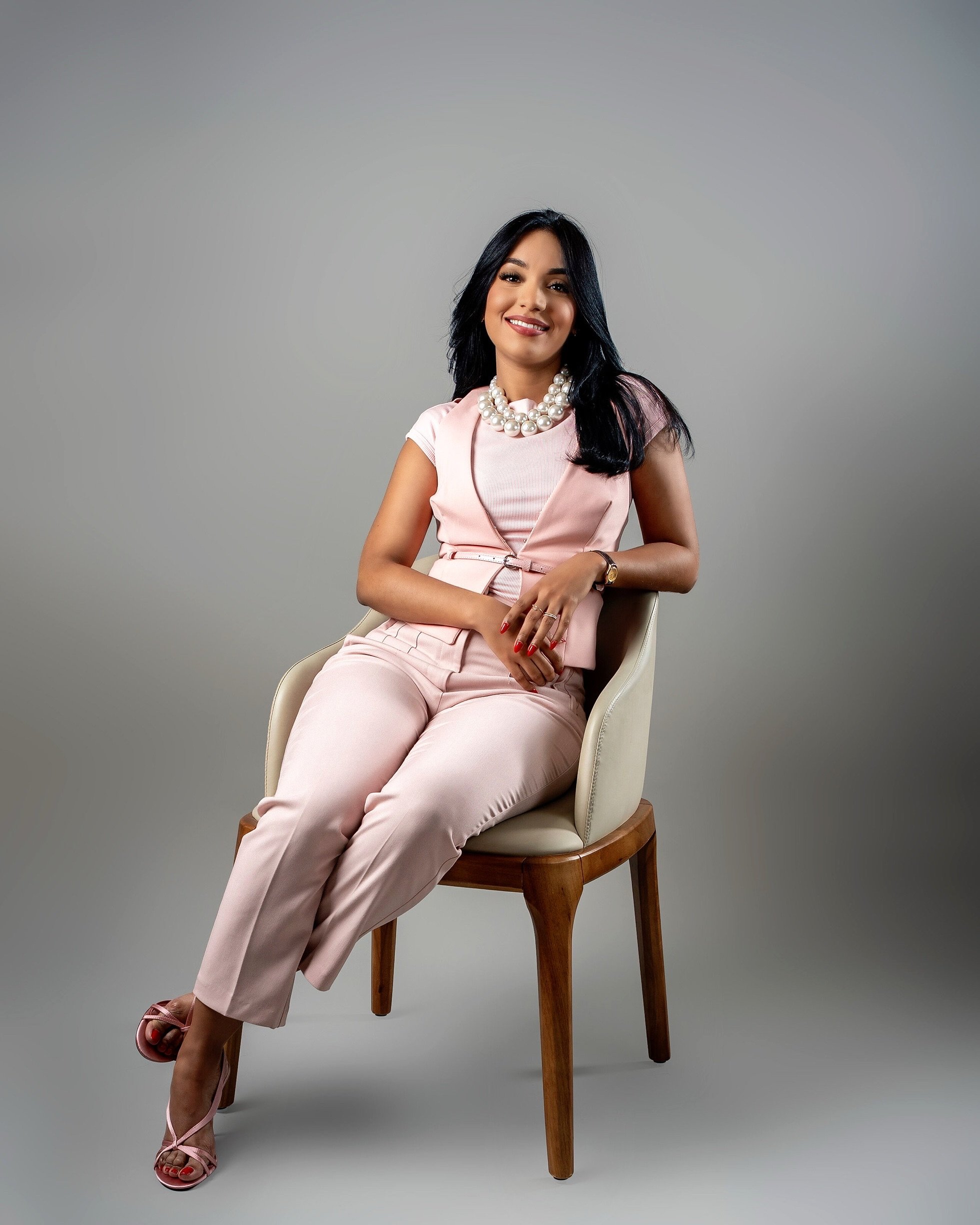 Mujer con vestido rosa y collar de perlas, sentada en una silla de madera y terciopelo, sonriendo.
