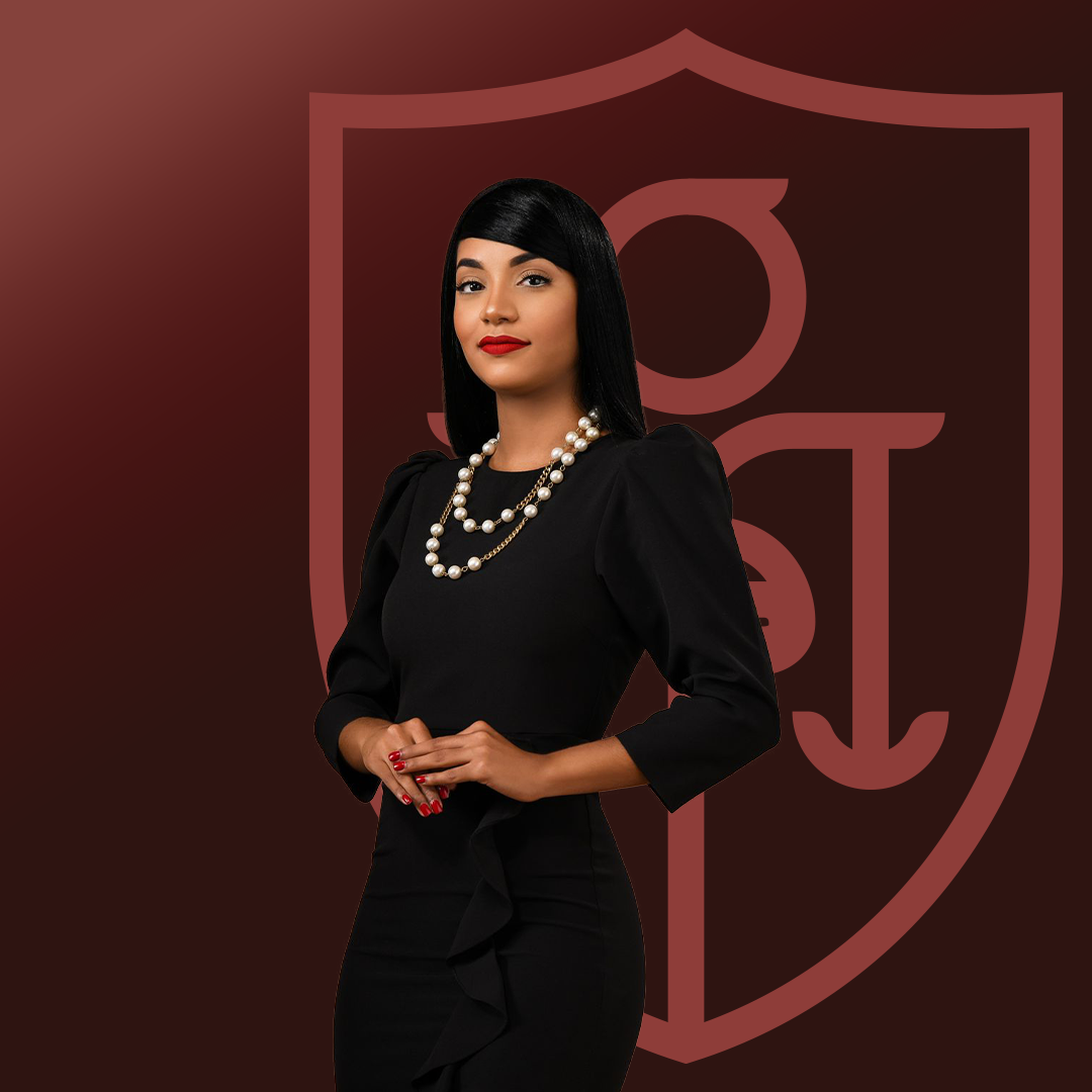 Mujer vestida con ropa elegante negra, con collar de perlas, de cabello negro y peinado liso, posando frente a un fondo con escudo en tonos burdeos.