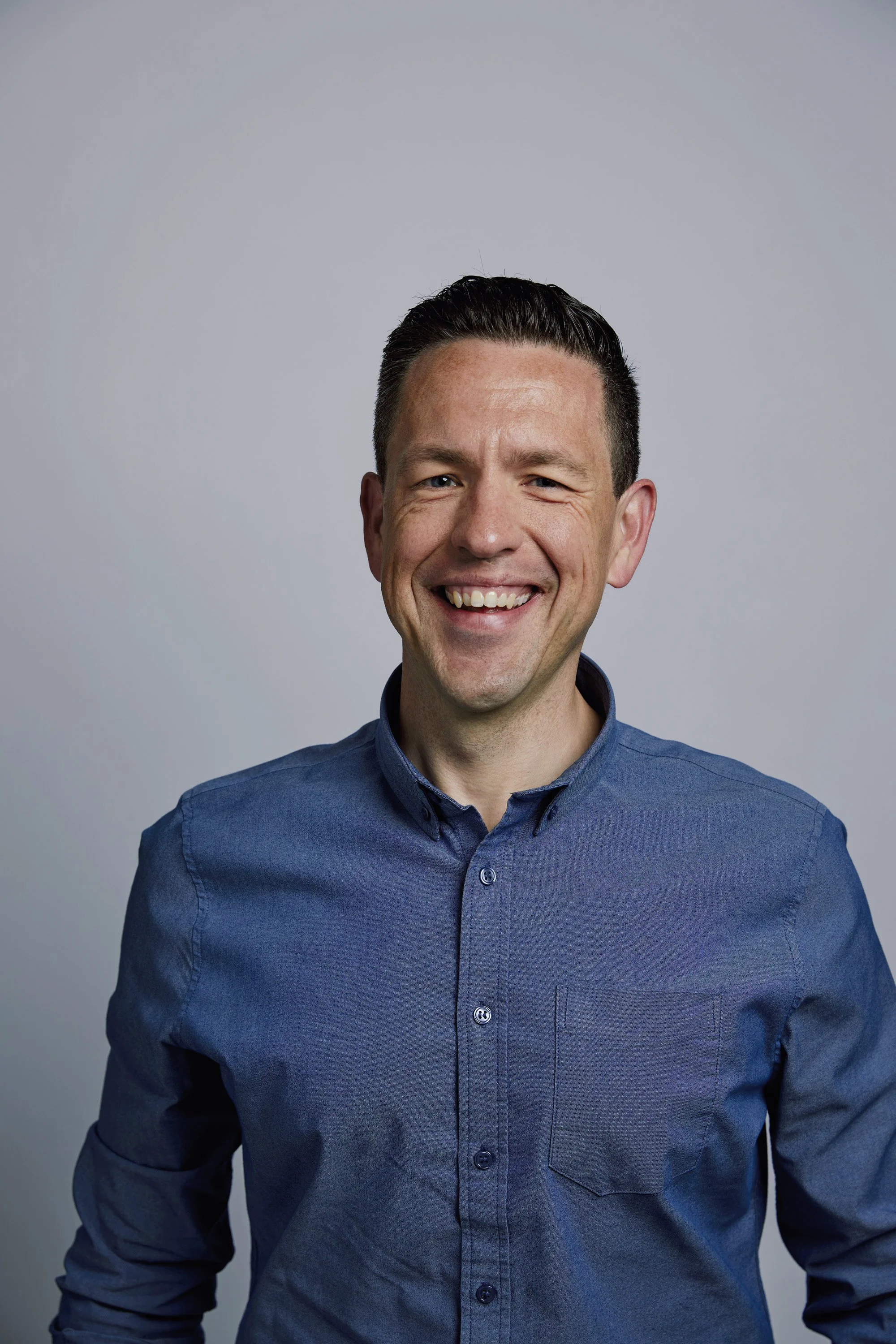 Headshot of a smiling man wearing a blue button-up shirt with a plain light gray background.