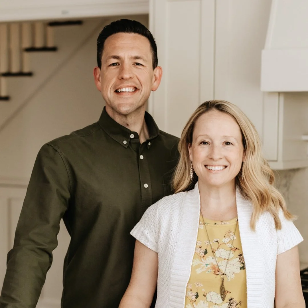 A man and woman smiling indoors with a neutral background and stairs.