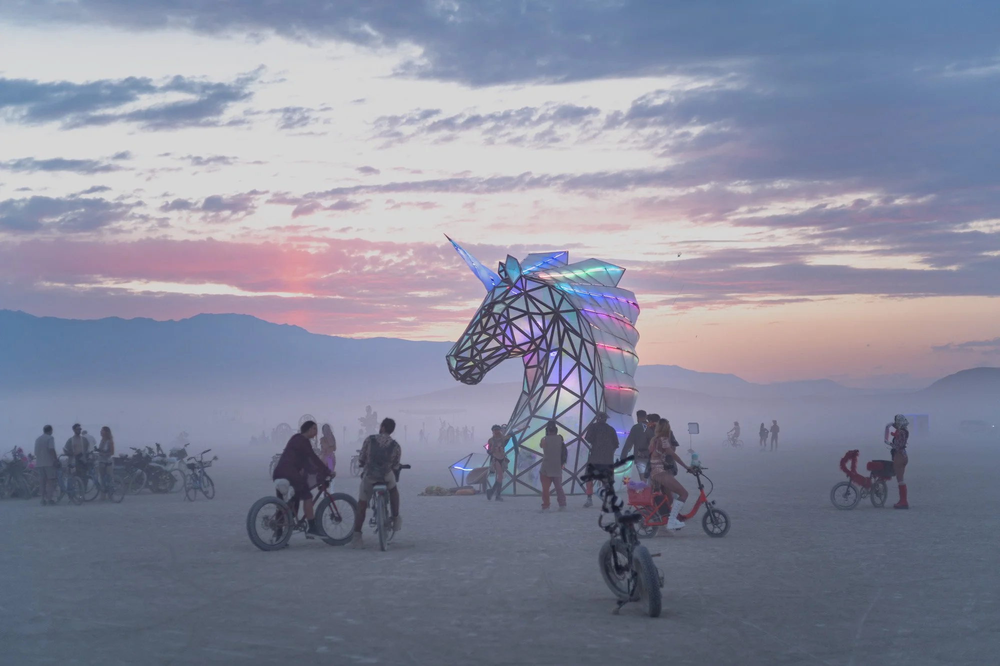 A large illuminated geometric unicorn sculpture with a rainbow-colored mane and horn stands in a desert during sunset or sunrise. People, including families and children riding bikes, are gathered around the sculpture in a dusty or misty environment with mountains in the background.
