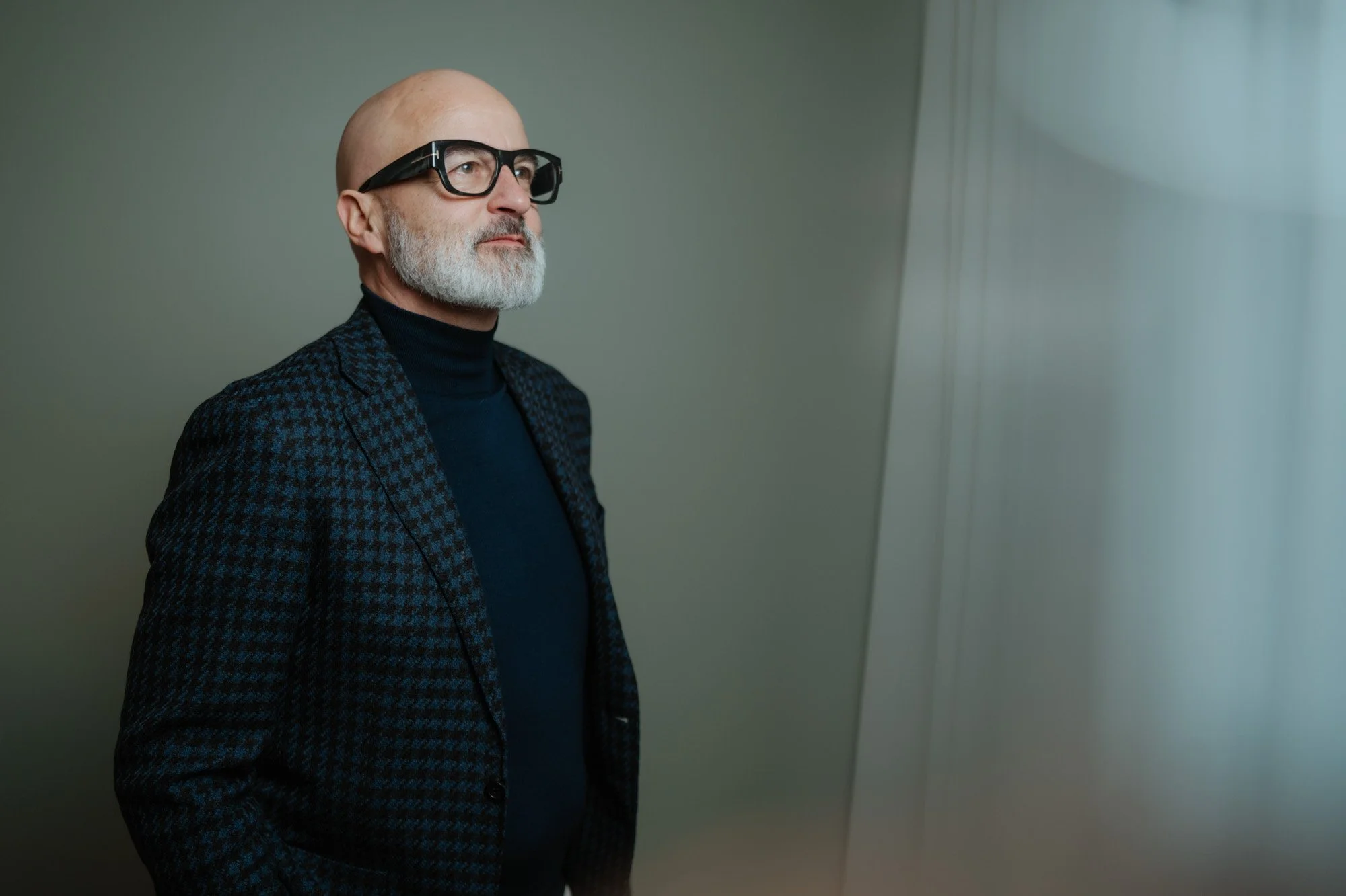 A bald man with a gray beard wearing glasses, a dark turtleneck, and a checkered blazer looking to the side.