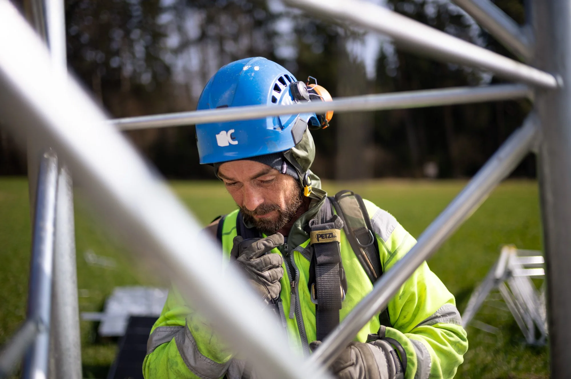 A man wearing a blue safety helmet, yellow high-visibility jacket, and gloves working on a metal structure outdoors in a park or field.