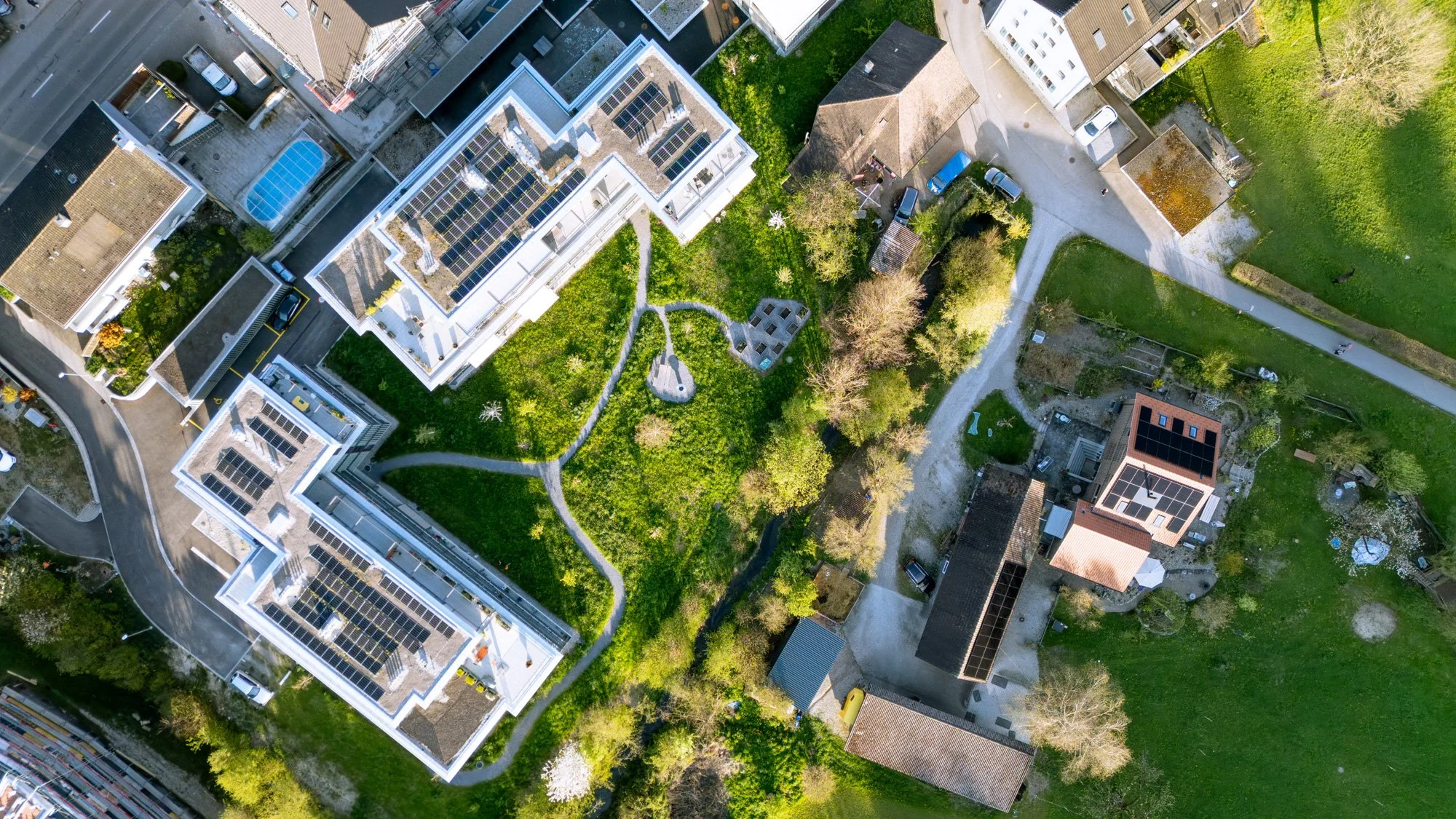 Aerial view of a city block showing green lawns, trees, white modern buildings with solar panels, parking lots, and surrounding streets.