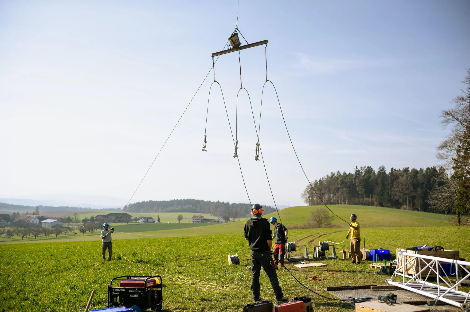 People working on assembling a large outdoor structure with a crane lifting a long support beam, set in a green field with trees and houses in the background.
