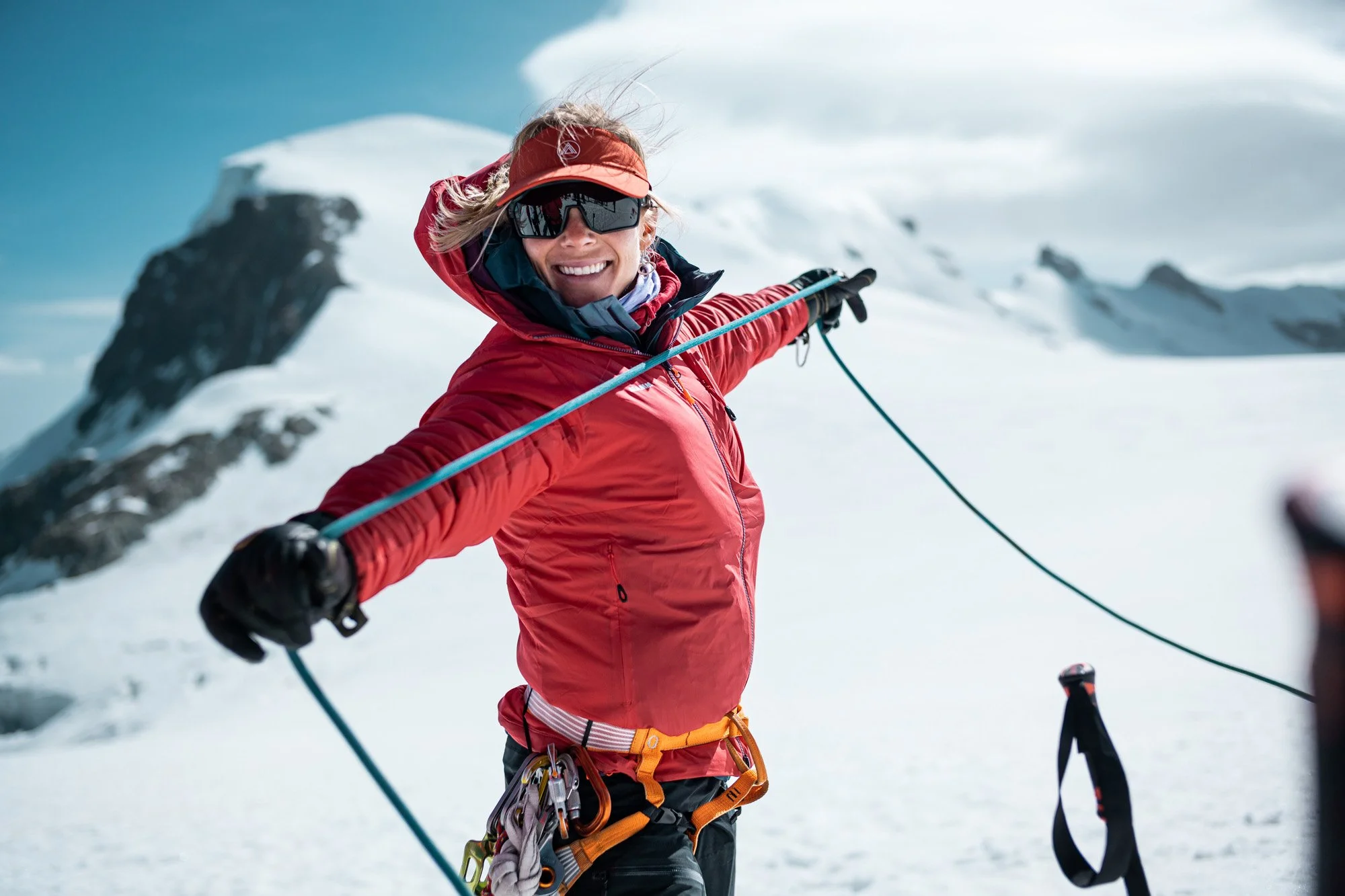 A smiling woman in a red jacket, sunglasses, and a red visor hat is taking a selfie on a snowy mountain, holding ski poles, with snow-covered peaks in the background.