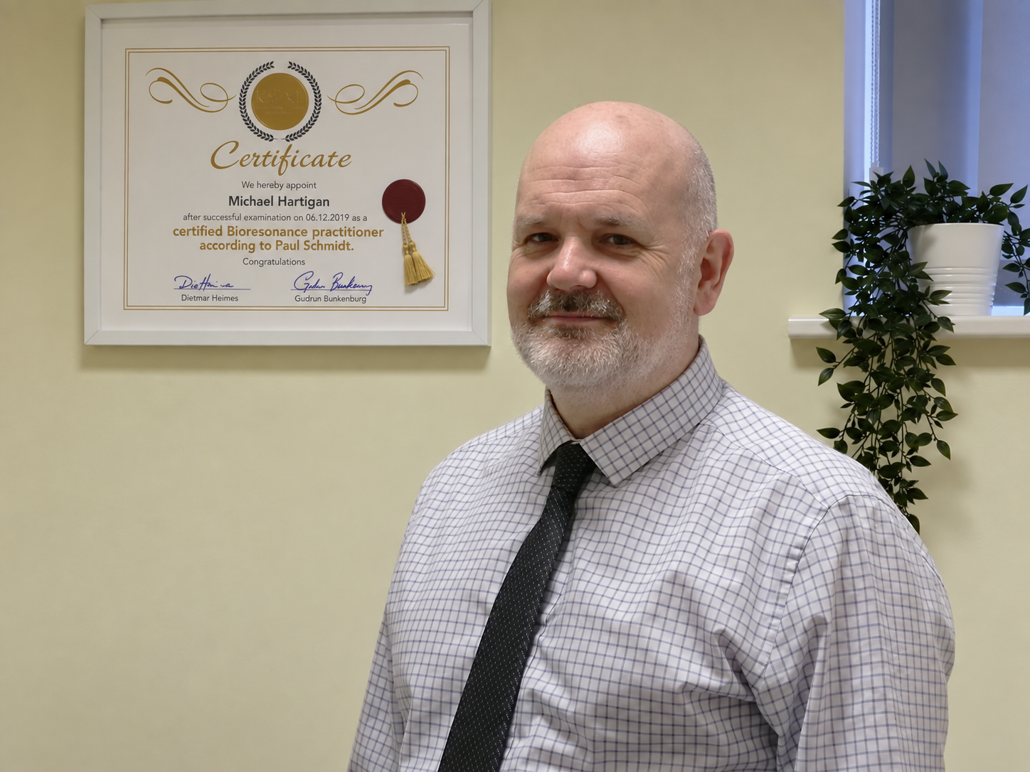 Man with short gray hair, beard, wearing a checkered shirt and dark tie, standing in front of a framed certificate on a beige wall, with a window and potted plant in the background.
