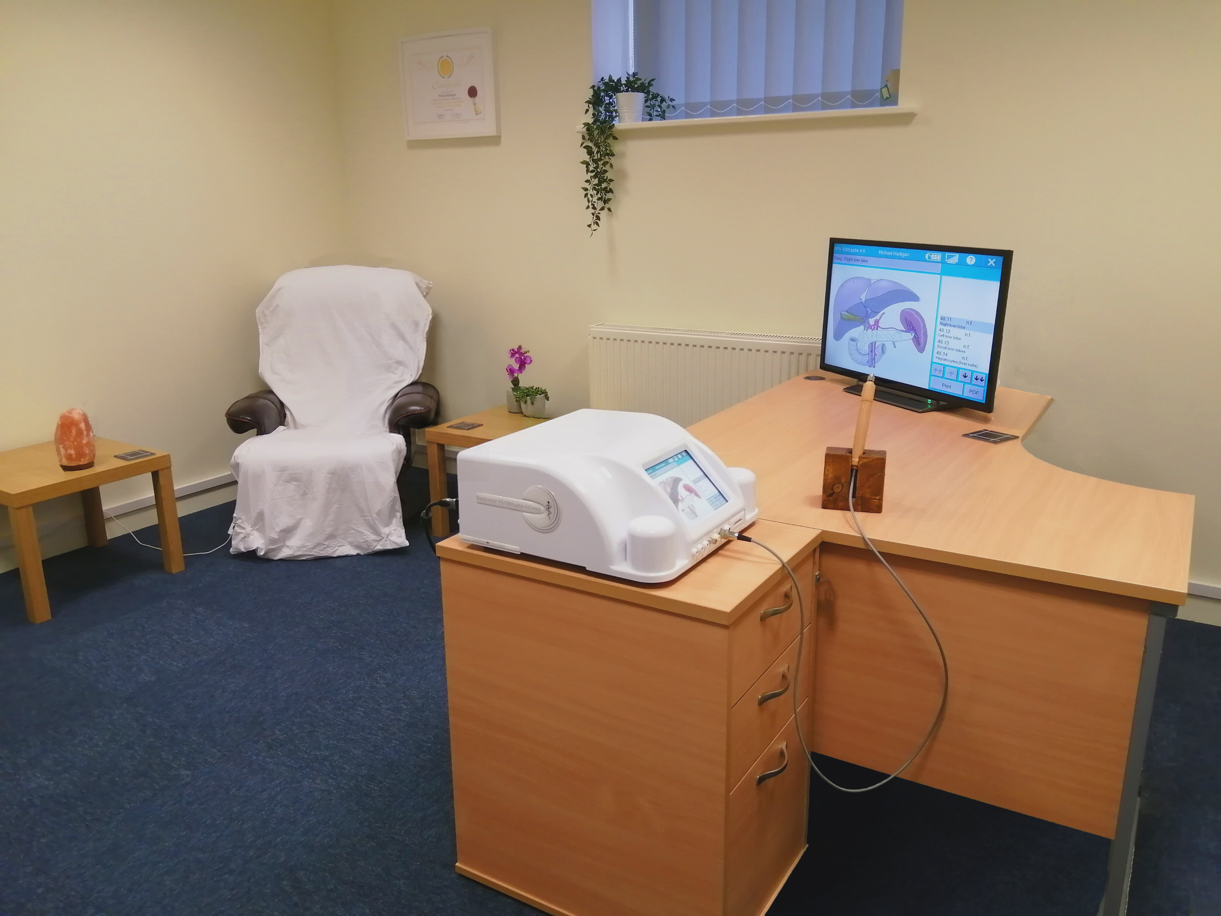 A medical room with a desk, computer monitor, treatment equipment, a white covered chair, side table with a salt lamp and potted plant, and medical certificates on the wall.
