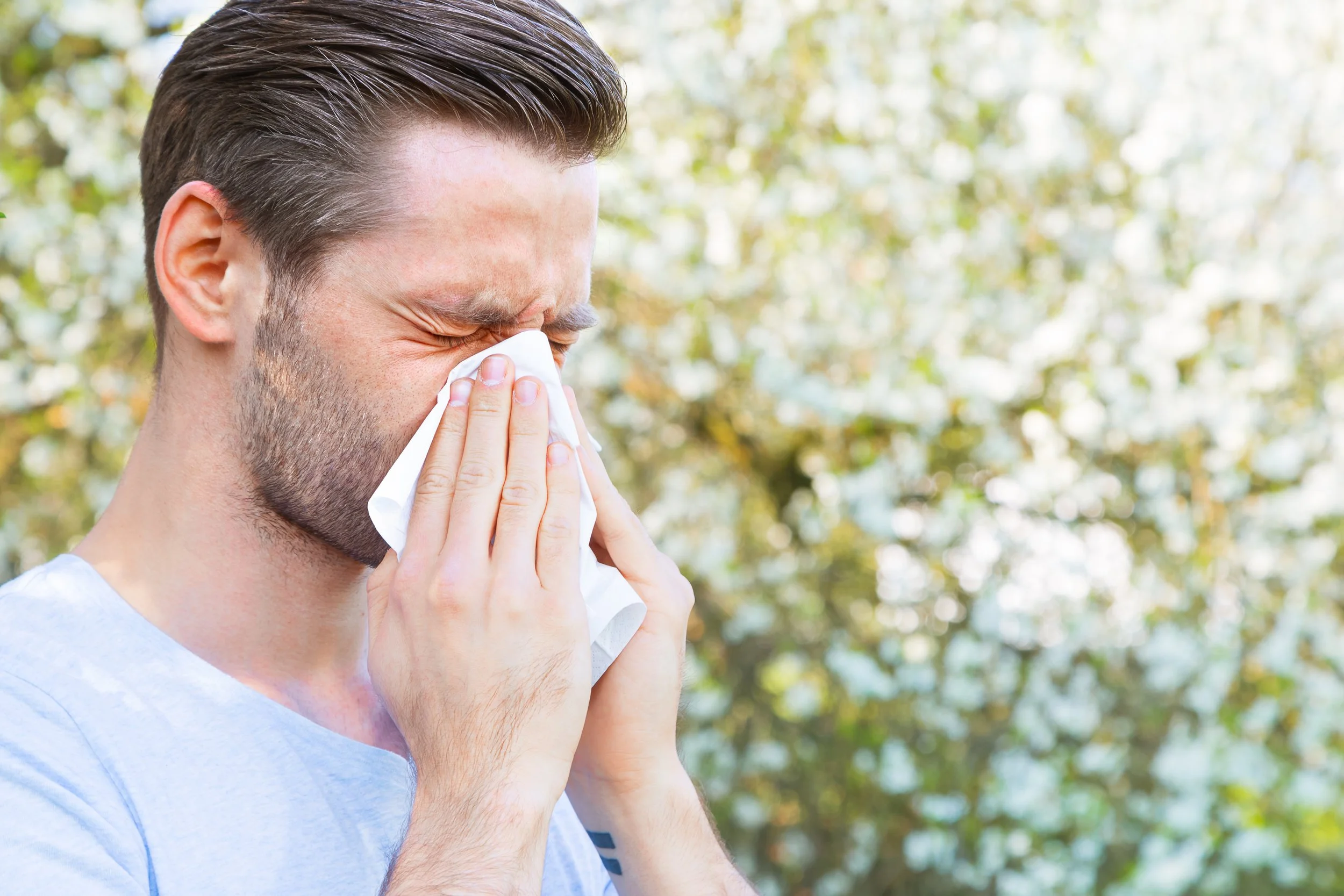 A man with brown hair and a beard, wearing a light blue shirt, is outdoors and appears to be sneezing or blowing his nose into a tissue.