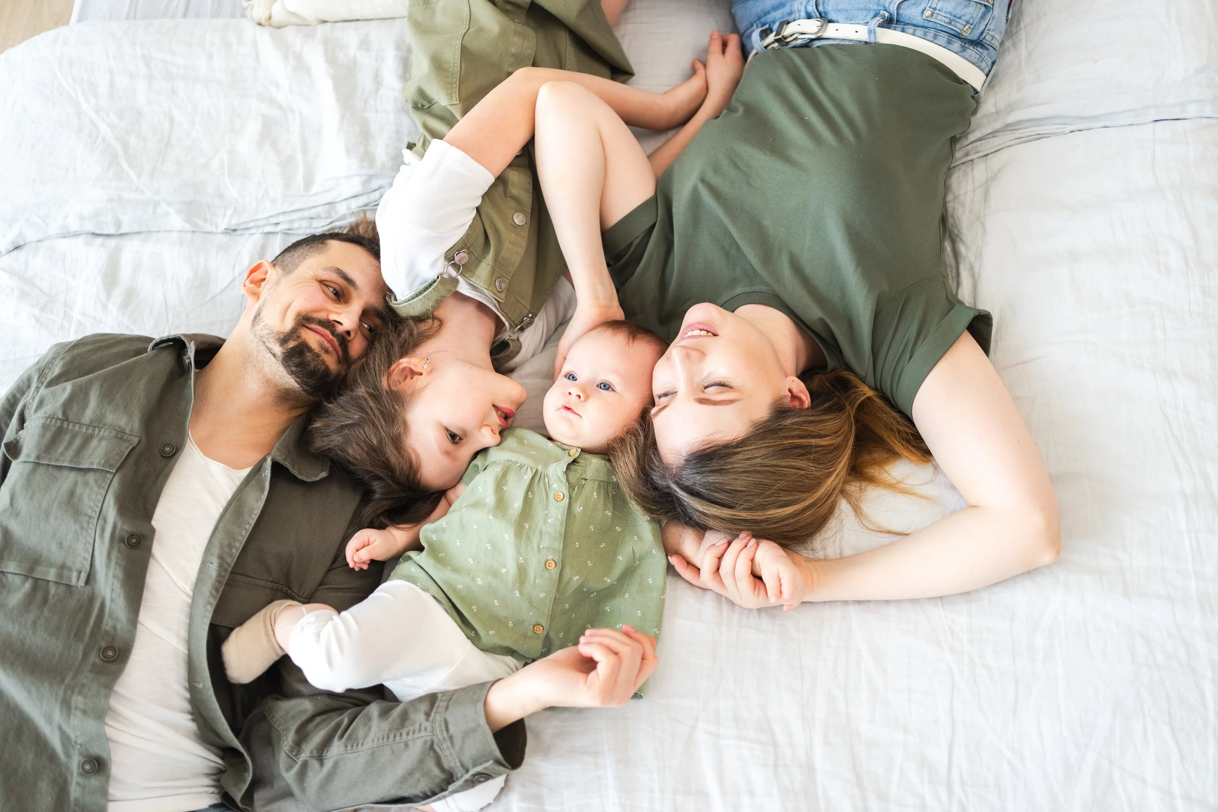 A family lying together on a bed, including a man with dark hair and beard, a woman with long hair, two young girls, and a baby, all relaxed and smiling
